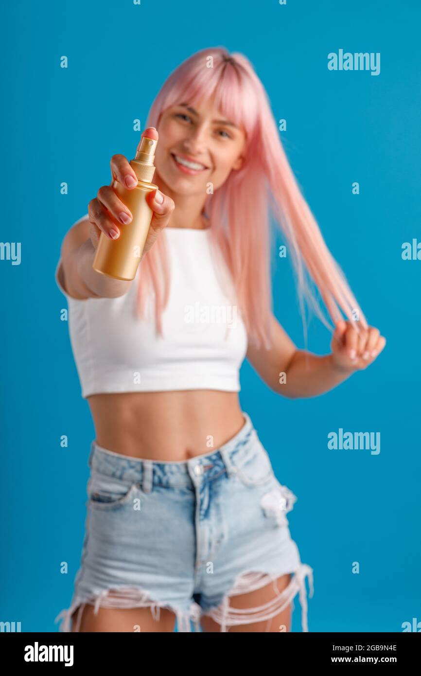 Giovane donna allegra con i capelli rosa sorridenti e che mostra spray idratante alla macchina fotografica mentre si trova isolato su sfondo blu studio Foto Stock