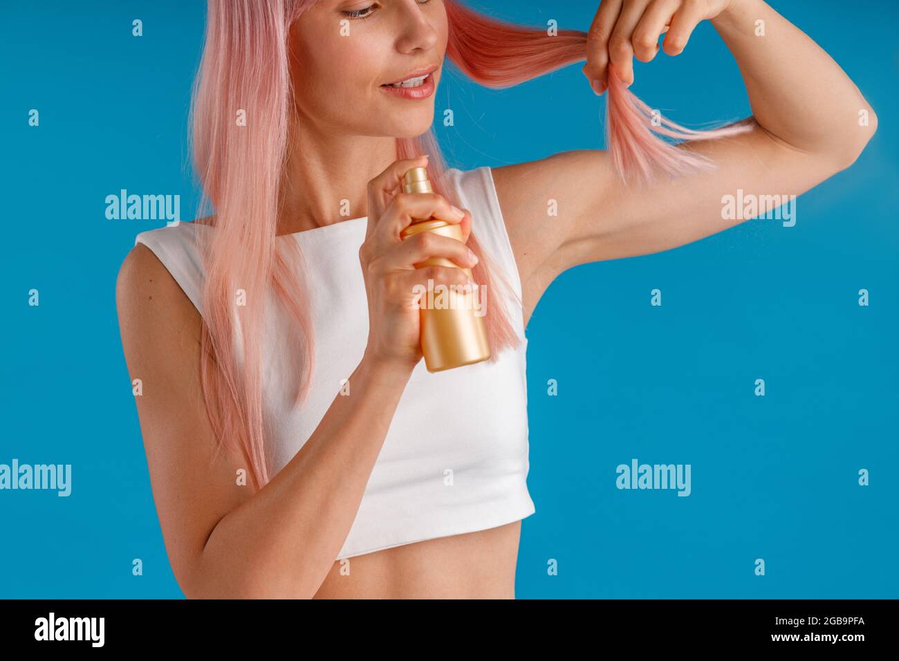 Primo piano di una giovane donna che applica spray idratante alle estremità dei capelli rosa mentre si trova isolato su sfondo blu studio Foto Stock