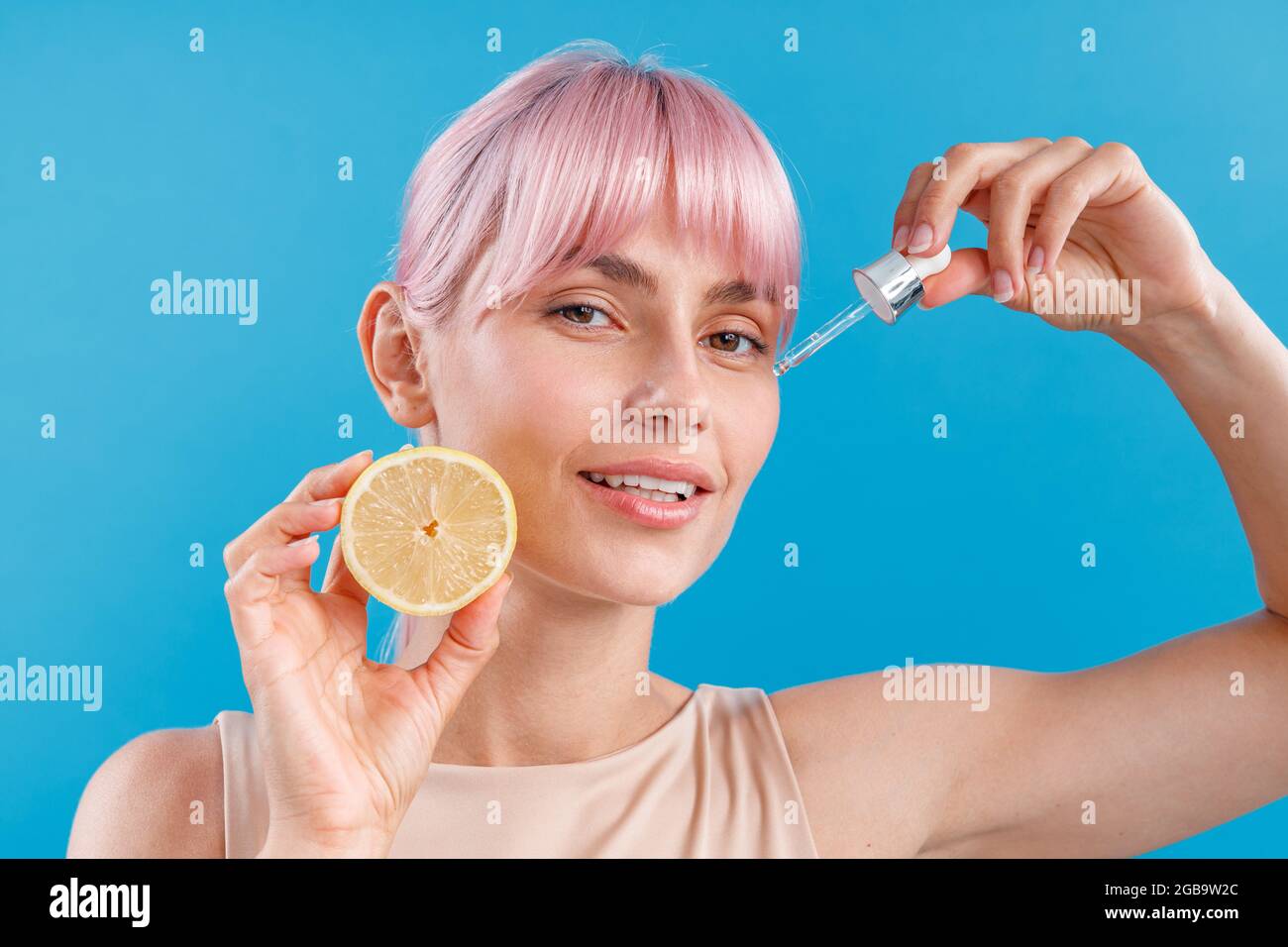 Ritratto di donna attraente con pelle perfetta e capelli rosa che tiene metà di limone fresco e un contagocce vicino al suo viso isolato su sfondo blu Foto Stock