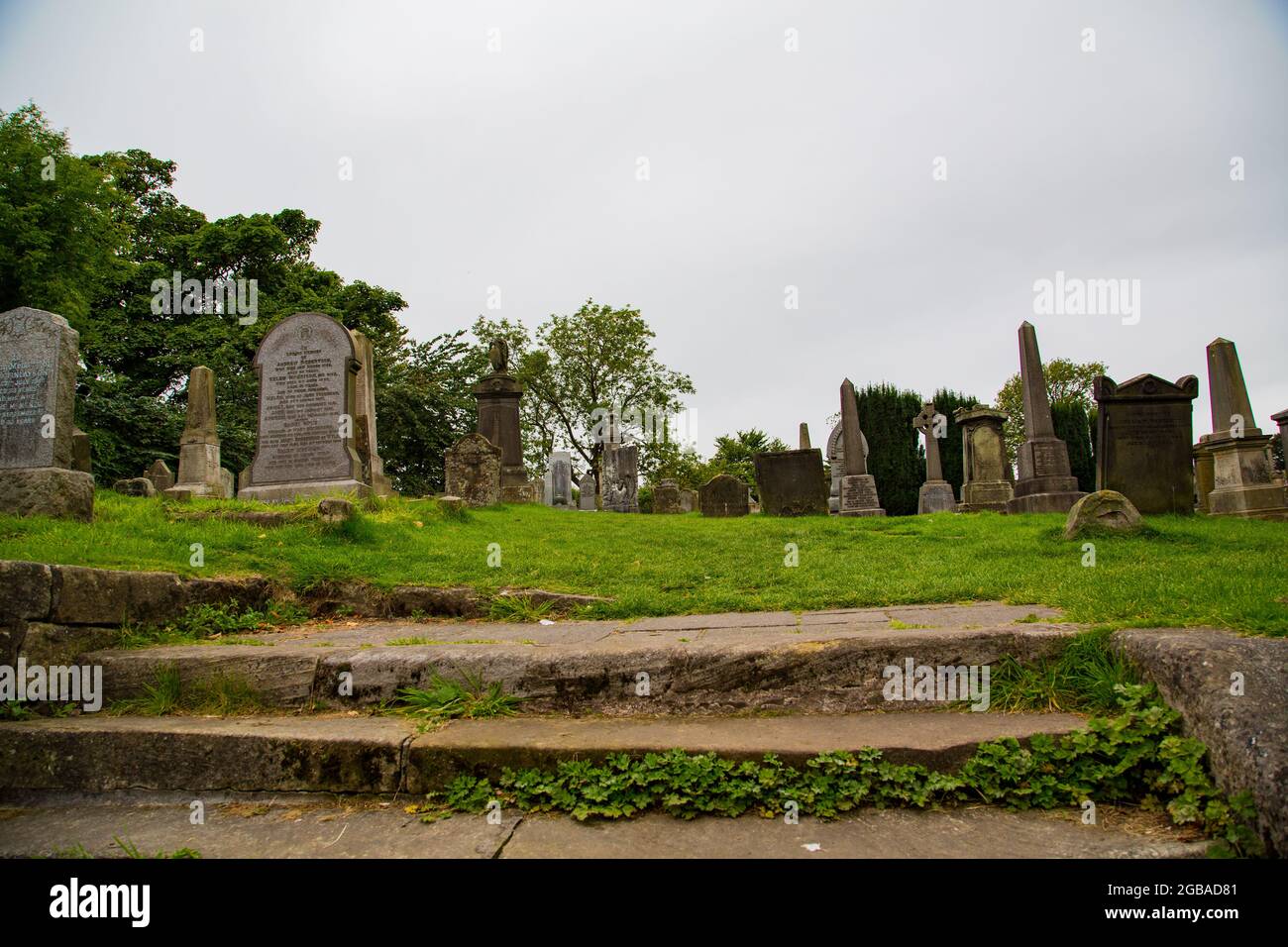 Cementerio de corte Gótico con lápidas en forma de piedra y monolitos, en prada con entrada de barrotes de hierro e iglesia detrás. Foto Stock