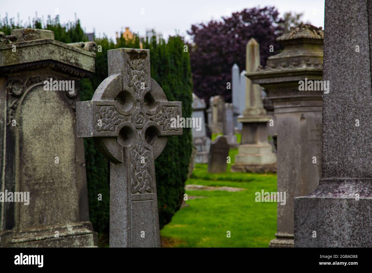 Cementerio de corte Gótico con lápidas en forma de piedra y monolitos, en prada con entrada de barrotes de hierro e iglesia detrás. Foto Stock