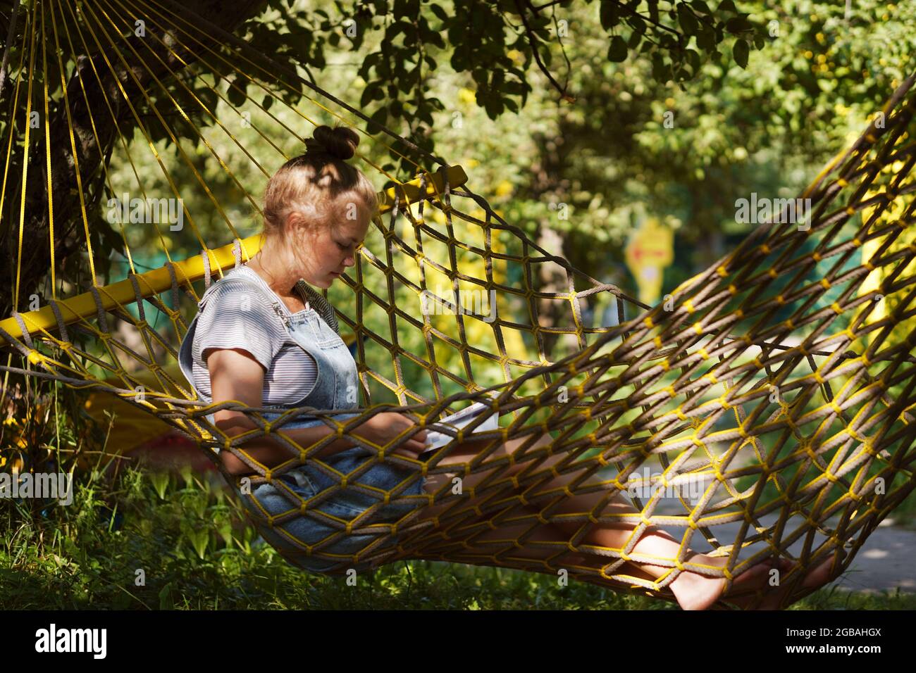 Giovane studentessa sorridente a leggere un libro mentre si steso su un'amaca in giardino. Donna che si rilassa nel libro di lettura amaca. Torna all'università, all'università Foto Stock