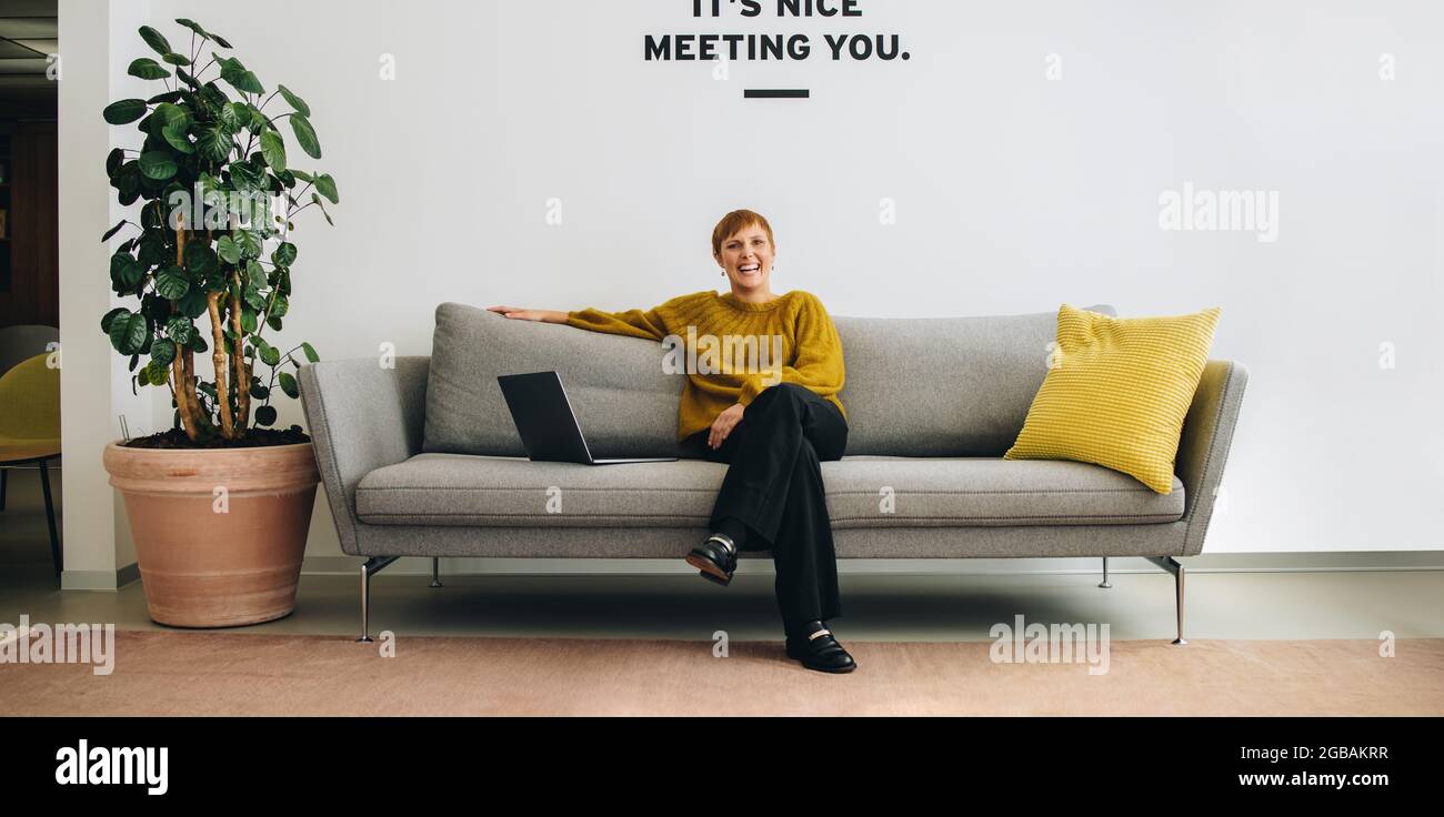 Una donna d'affari sorridente che si siede sul divano nella lobby dell'ufficio. Imprenditore allegro sul divano. Foto Stock