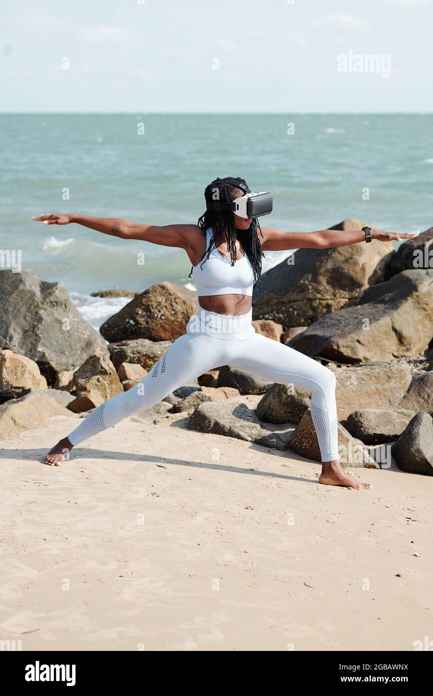 Indossa una giovane donna che indossa occhiali di realtà virtuale quando pratichi lo yoga sulla spiaggia e fai la posa del guerriero Foto Stock