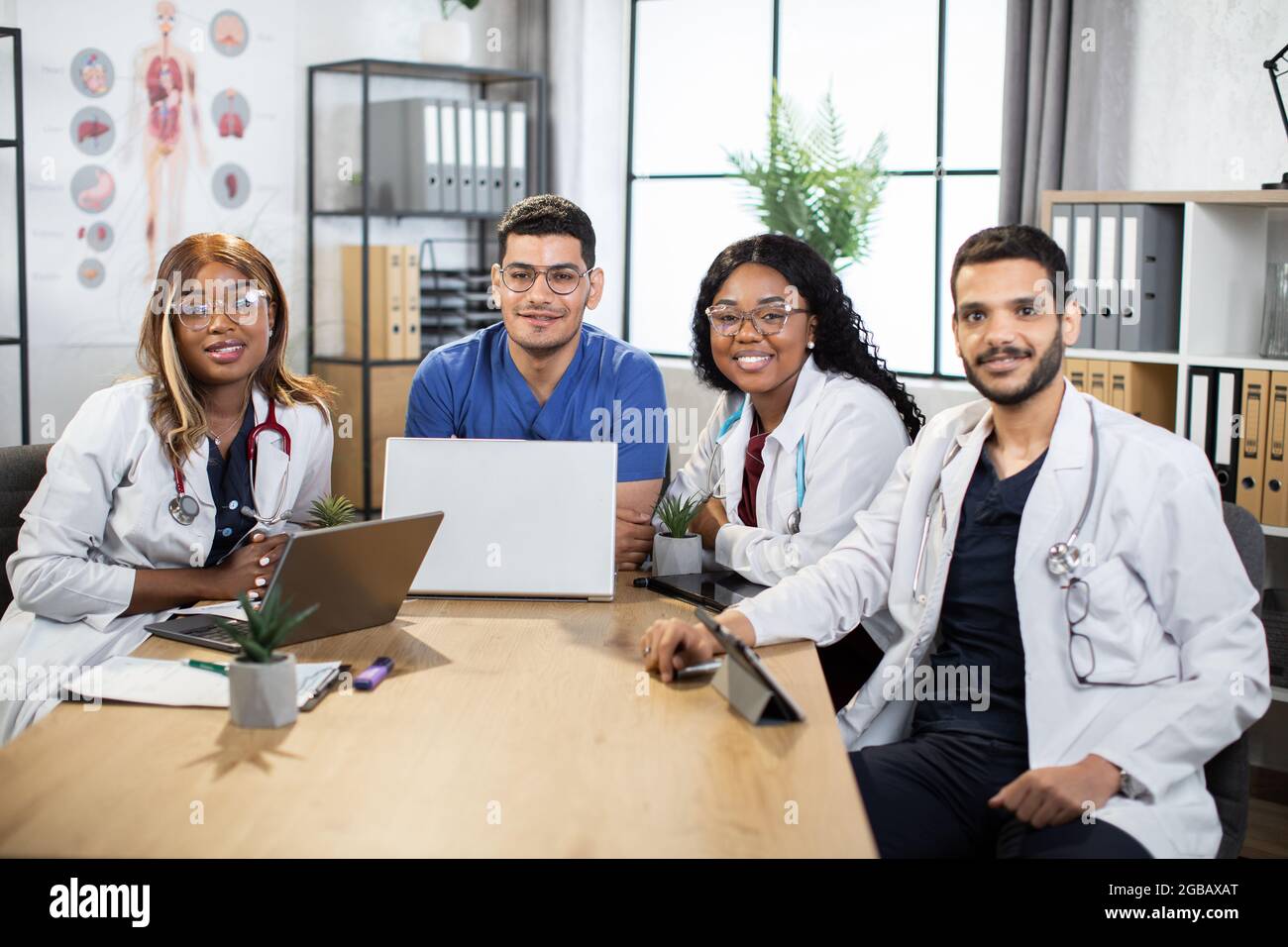 Due uomini arabi e due donne afro-americane scienziati medici, seduti a tavola e utilizzando gadget moderni. Medici multiraciali che lavorano insieme su un nuovo progetto scientifico, sorridendo alla telecamera Foto Stock