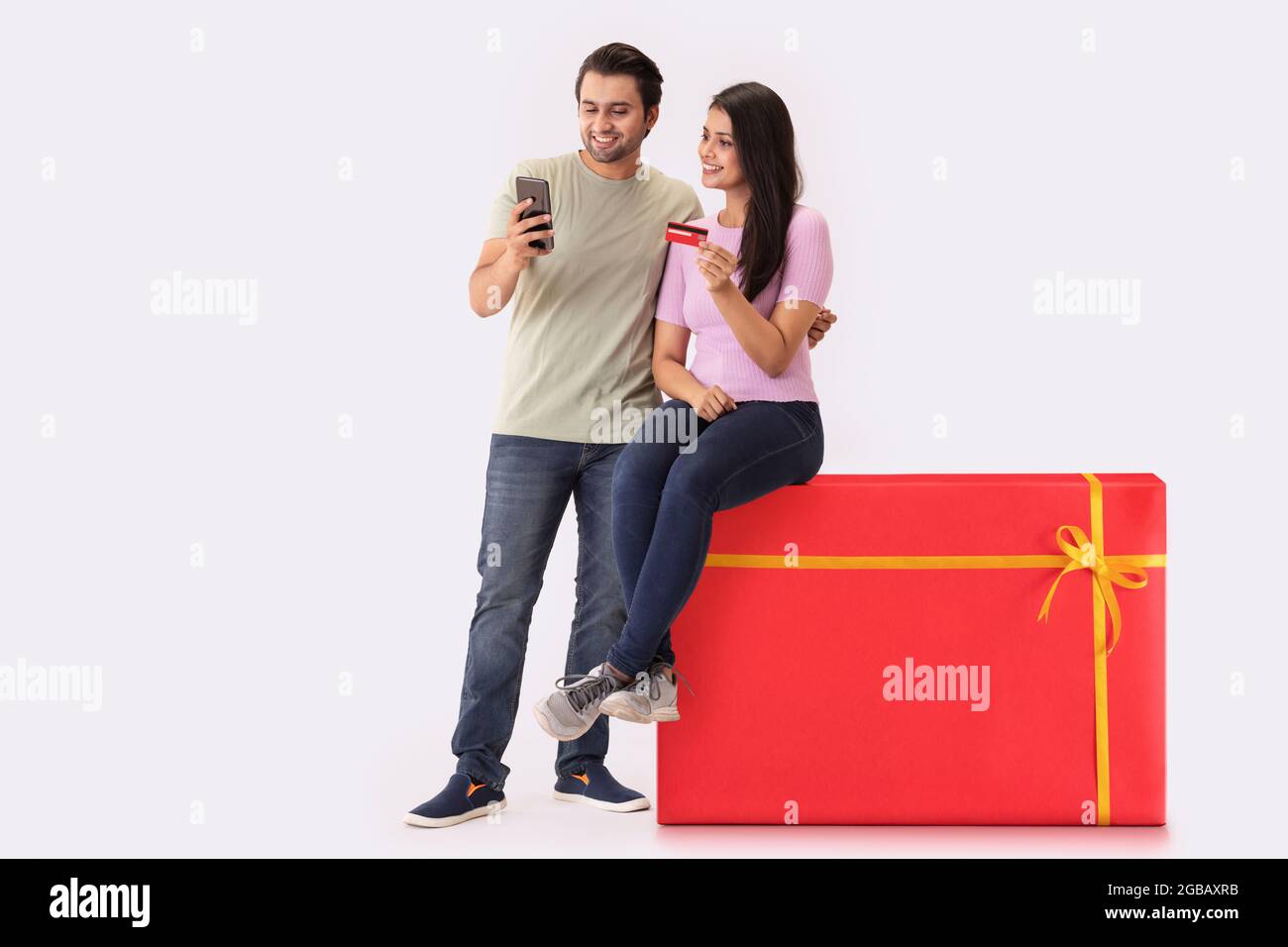 Una giovane donna seduta su uno sgabello a forma di regalo con un uomo accanto, facendo il pagamento online con carta di credito e telefono. Foto Stock