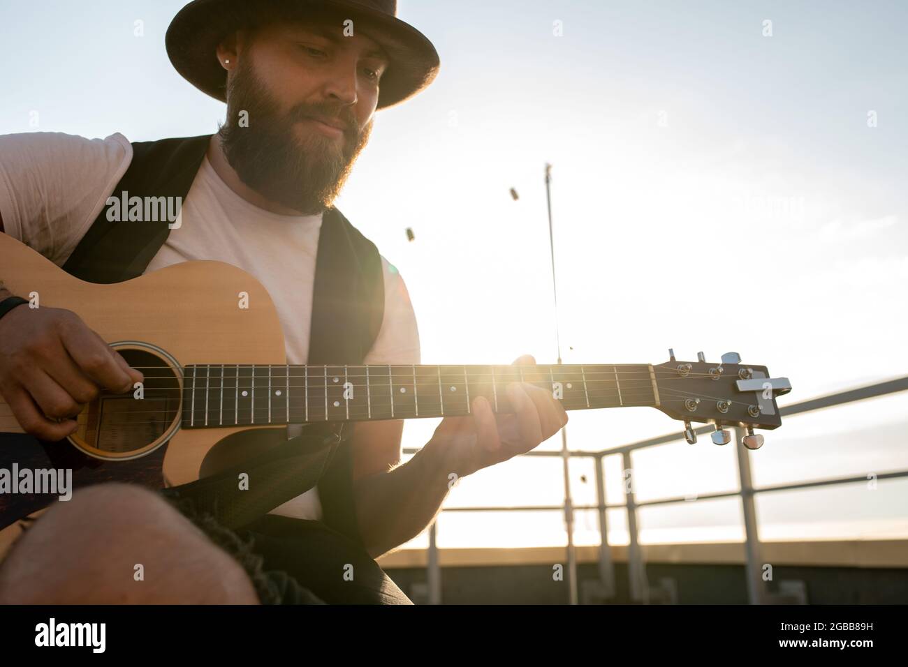 Giovane uomo bearded in cappello che suona la chitarra sul tetto il giorno di sole Foto Stock