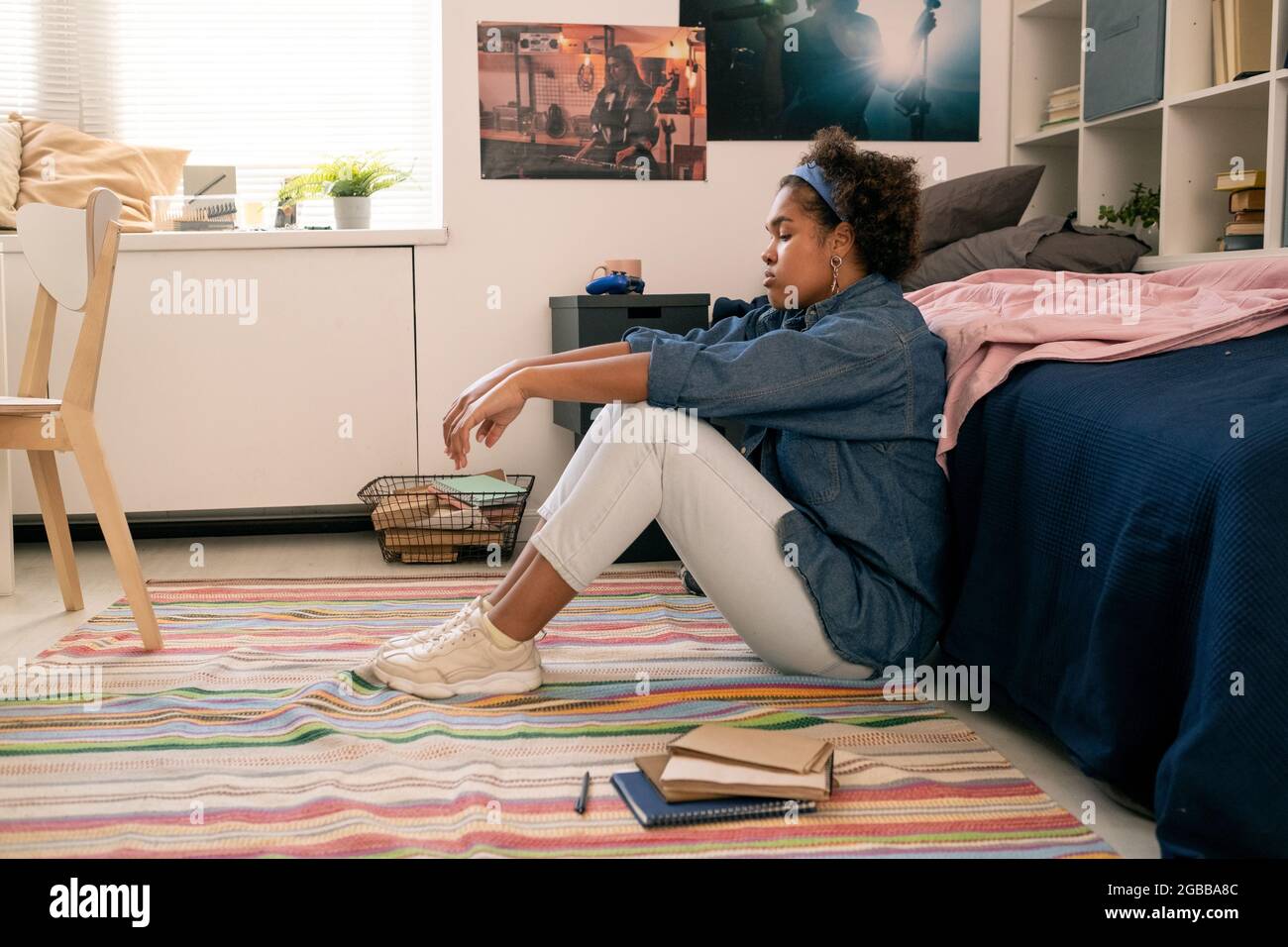 Stanca o turbata ragazza adolescente in casualwear seduta sul pavimento vicino al suo letto Foto Stock