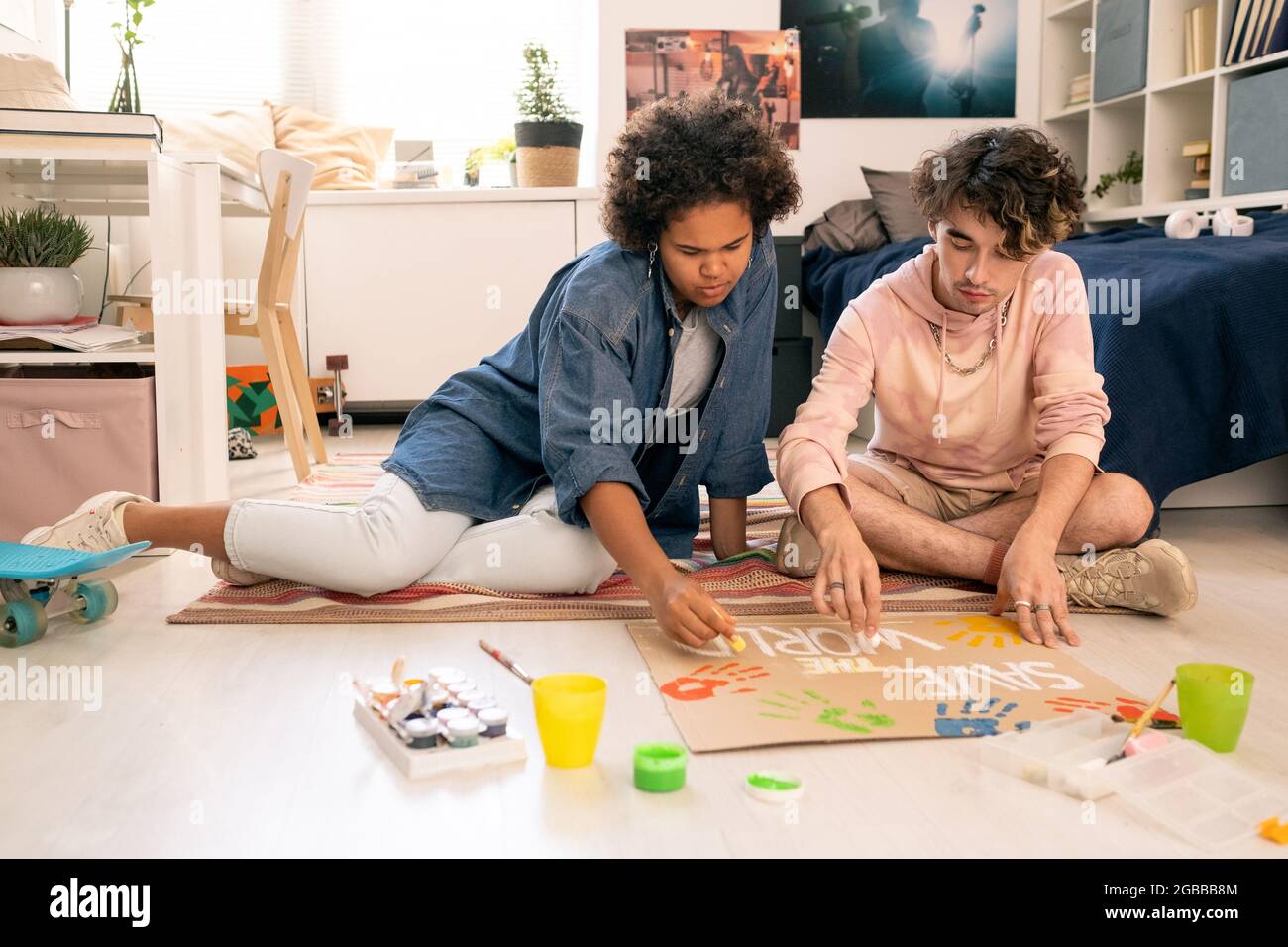 Cartello di disegno interculturale di coppia adolescente su pezzo di cartone mentre si siede sul pavimento Foto Stock