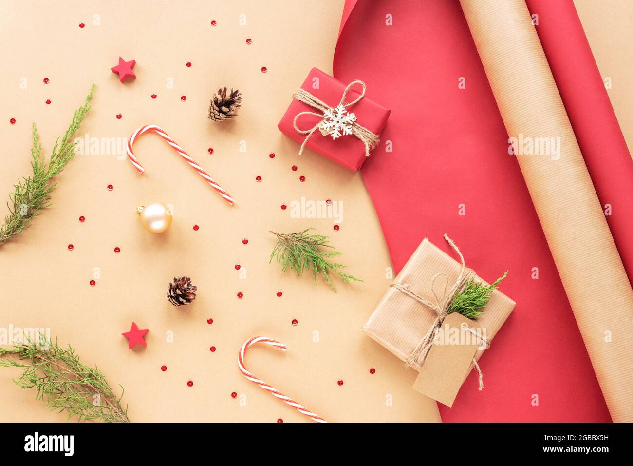 Scatole regalo, canne caramelle e rami di cipresso. Preparazione di regali di Natale su carta da imballaggio rossa e beige. Concetto di nuovo anno. Vista dall'alto, disposizione piatta. Foto Stock