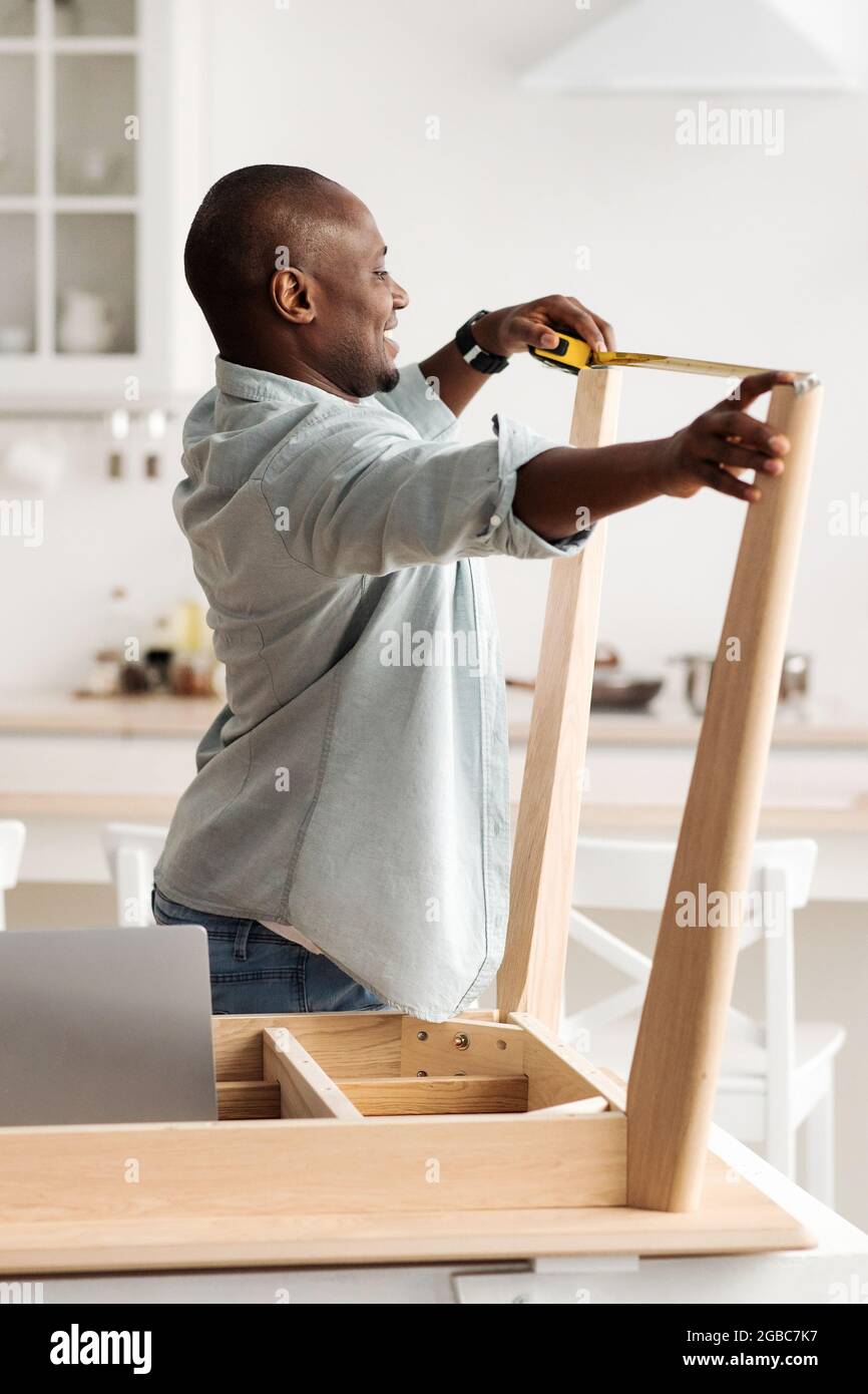 Manipolo millenario nero assemblando tavolo in legno e misurando la larghezza tra le gambe, in piedi in cucina, raccolto Foto Stock