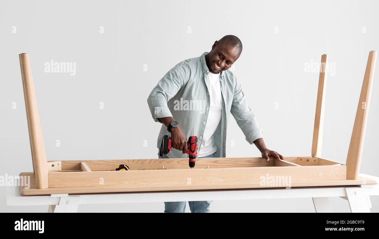 Happy handyman afro-americano forare tavole di legno di tavola, assemblando nuovi mobili con trapano elettrico, panorama Foto Stock