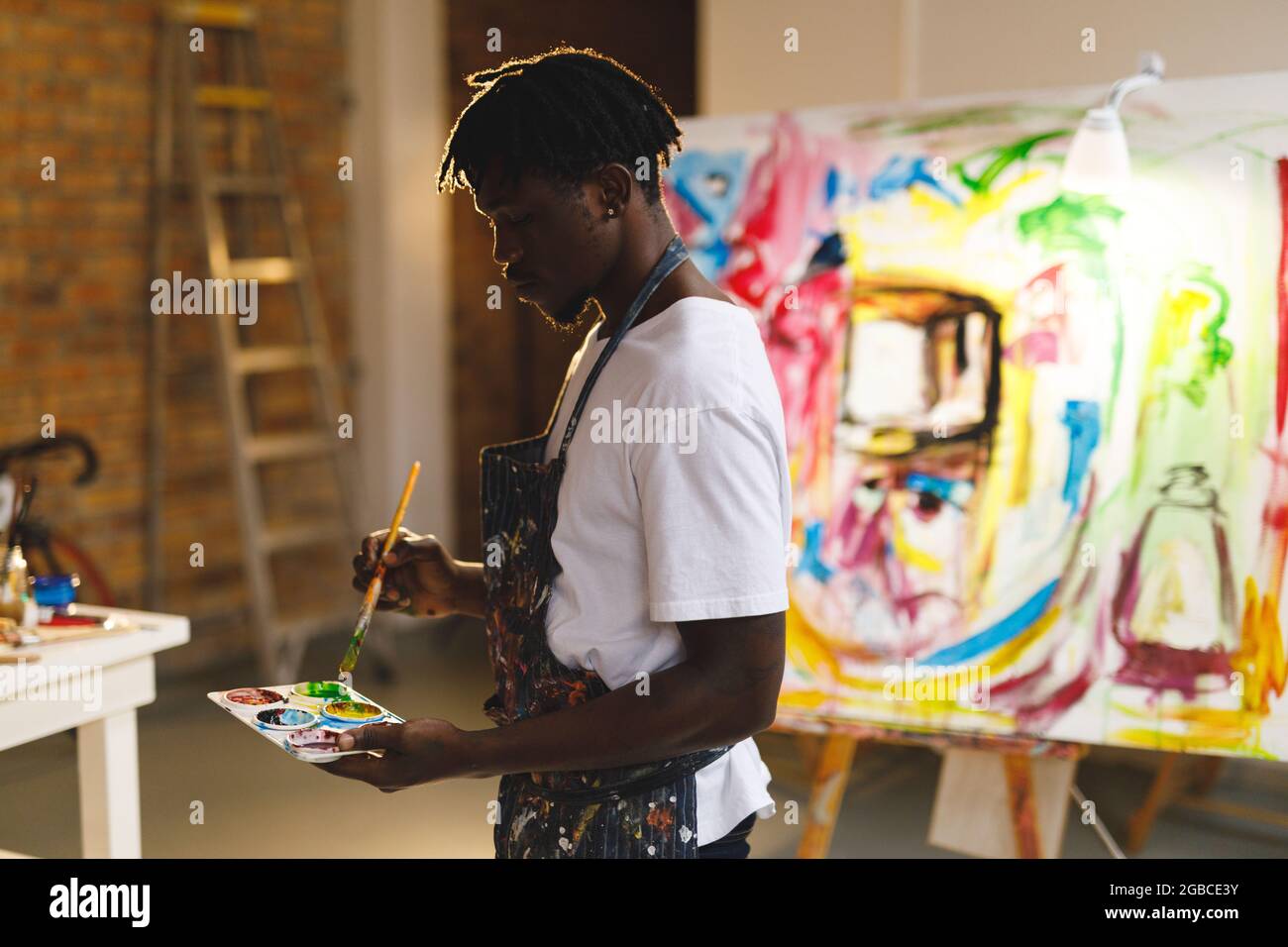 Pittore afroamericano maschio al lavoro che tiene vernici e pennellate in studio d'arte Foto Stock