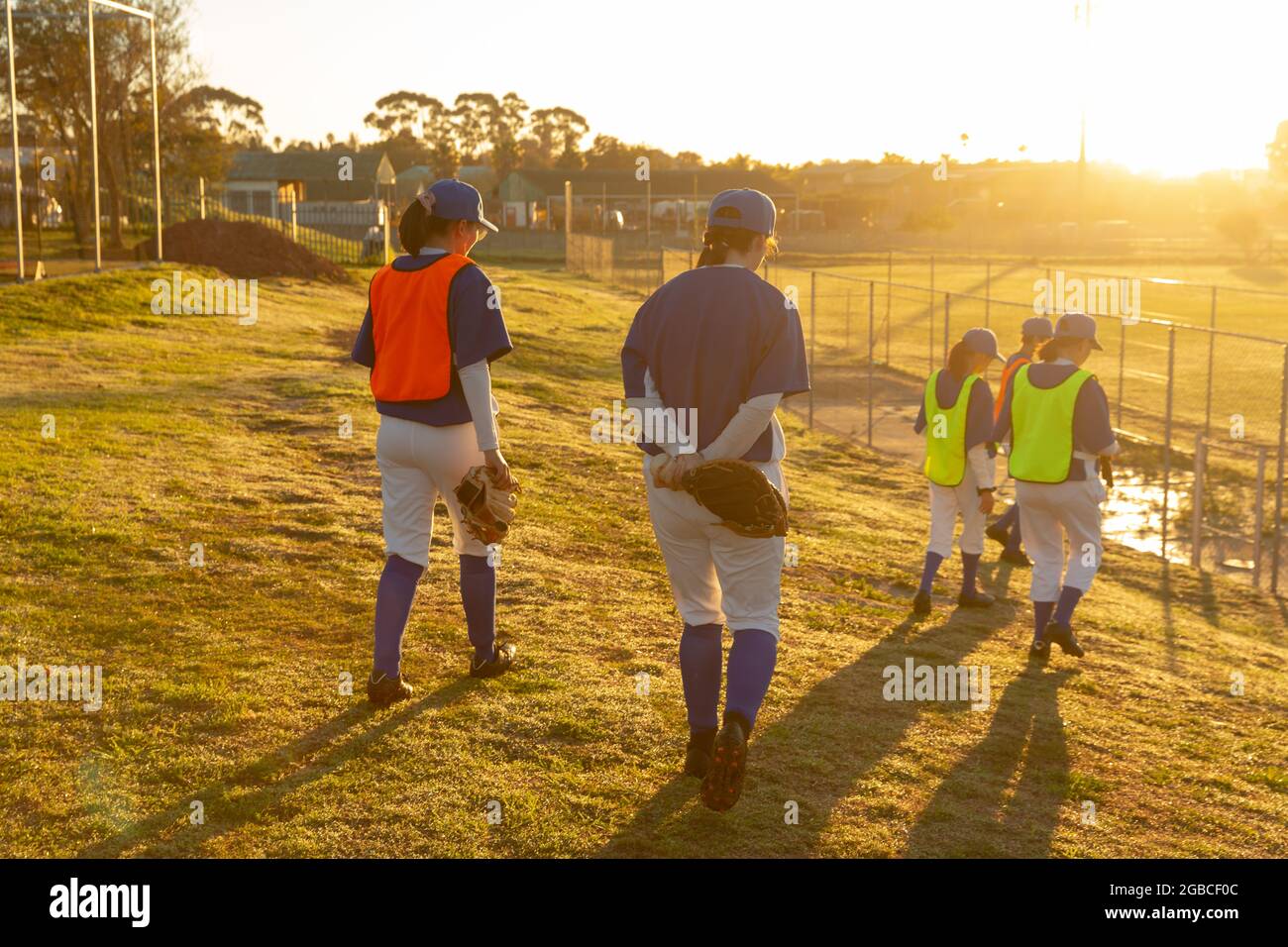 Un gruppo eterogeneo di giocatori di baseball femminili che camminano verso il campo all'alba per allenarsi Foto Stock