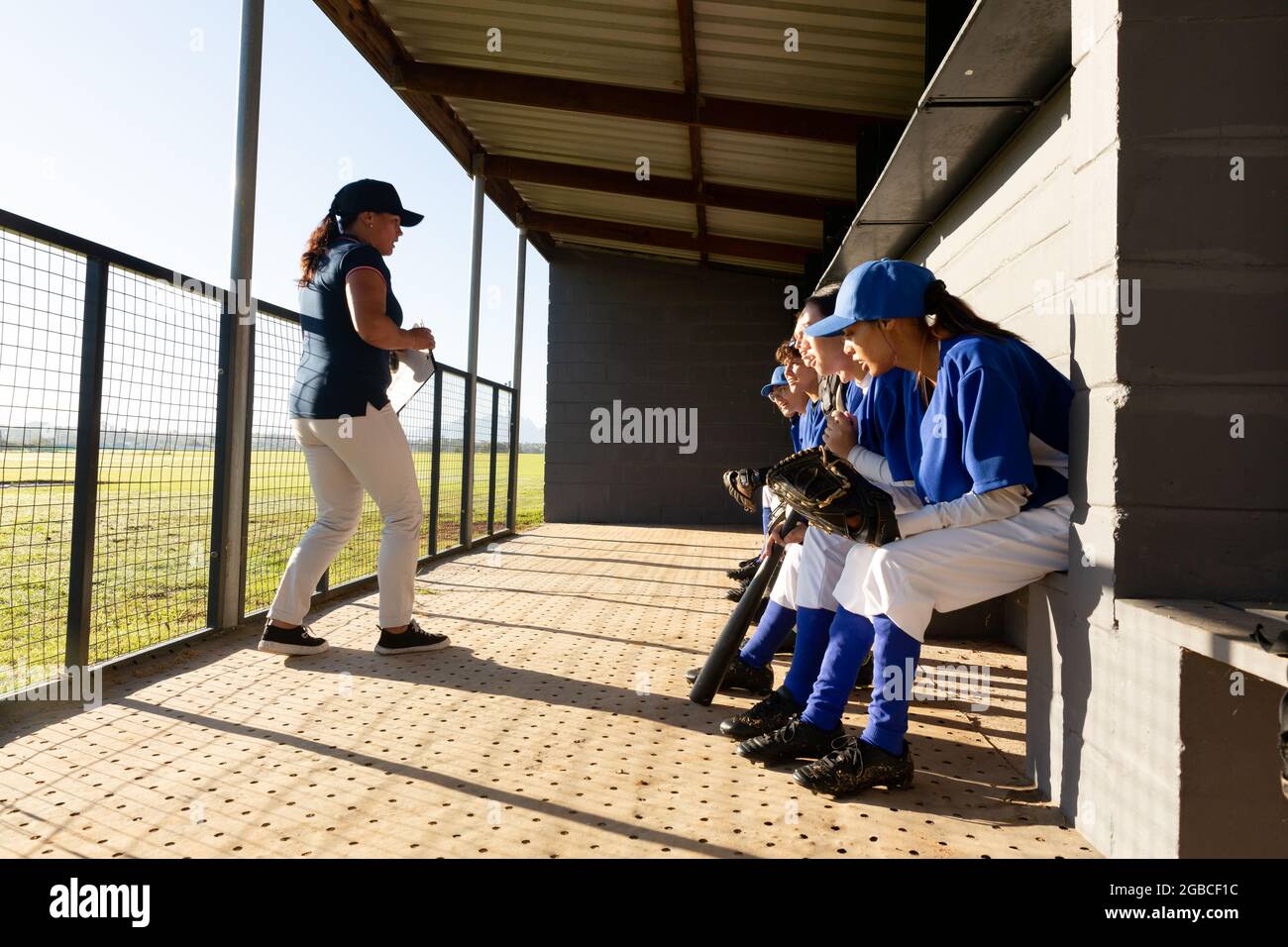 Un gruppo eterogeneo di giocatori di baseball femminili seduti sulla panchina, ascoltando la allenatrice femminile prima della partita Foto Stock