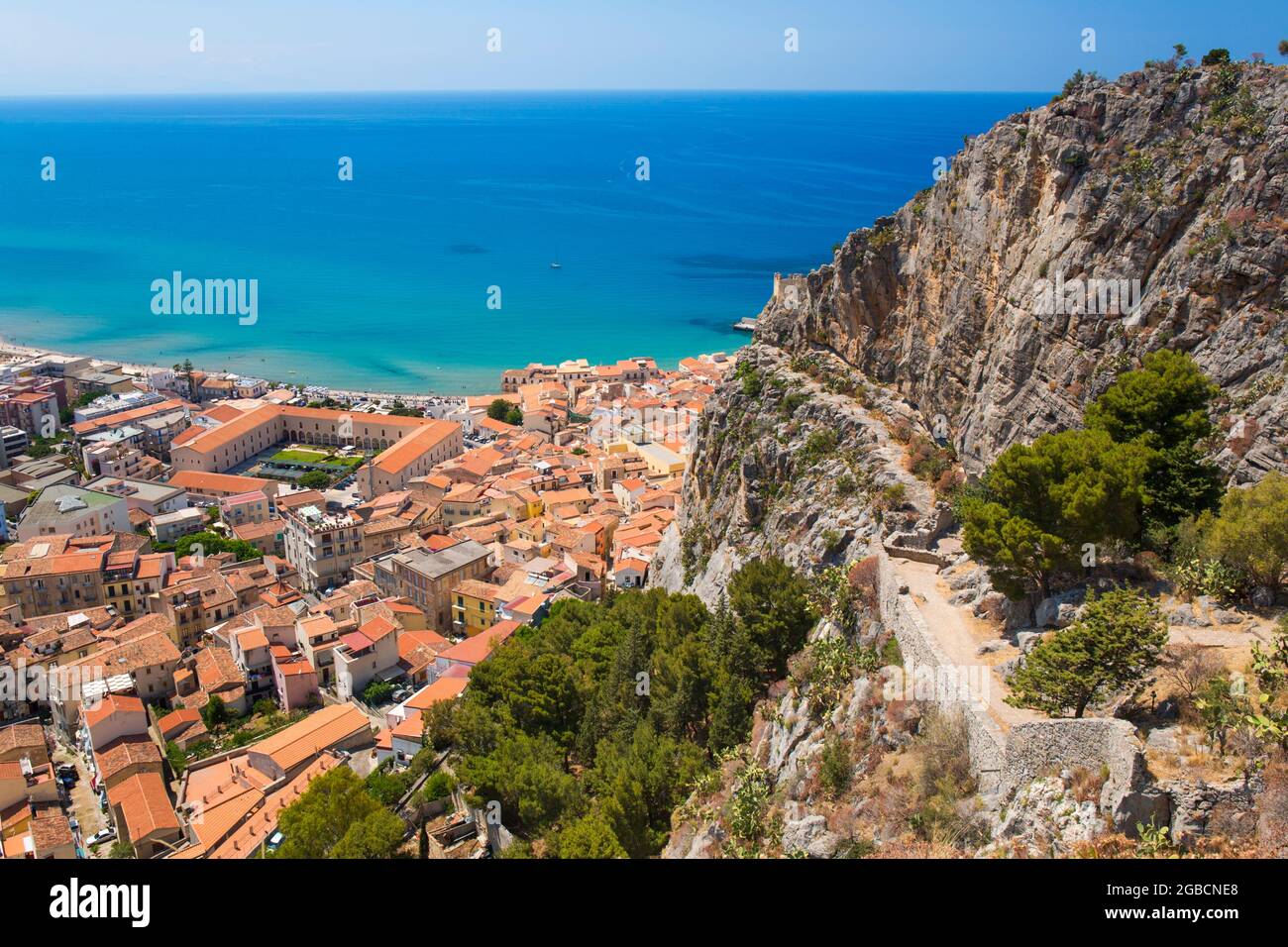 Cefalù, Palermo, Sicilia, Italia. Tetti della città dwarfed dalle torreggianti scogliere di la Rocca, le acque turchesi del Mar Tirreno oltre. Foto Stock