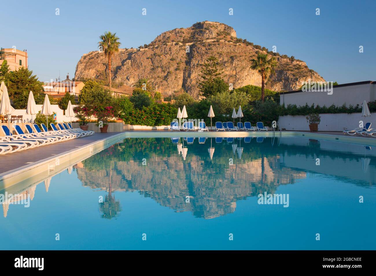 Cefalù, Palermo, Sicilia, Italia. Le torreggianti scogliere illuminate dal sole di la Rocca si riflettono nella tranquilla piscina dell'Hotel Kalura, alba. Foto Stock