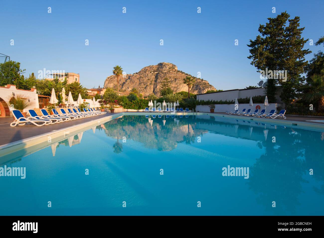 Cefalù, Palermo, Sicilia, Italia. Le torreggianti scogliere illuminate dal sole di la Rocca si riflettono nella tranquilla piscina dell'Hotel Kalura, alba. Foto Stock