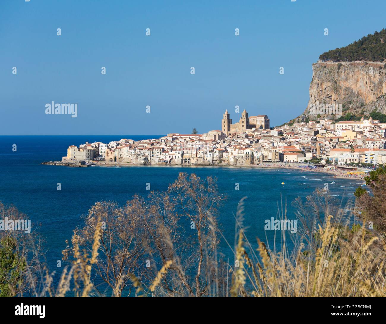 Cefalù, Palermo, Sicilia, Italia. Vista sulla baia dalla collina erbosa, la città e la cattedrale arabo-normanna dwarfed dalle torreggianti scogliere di la Rocca. Foto Stock