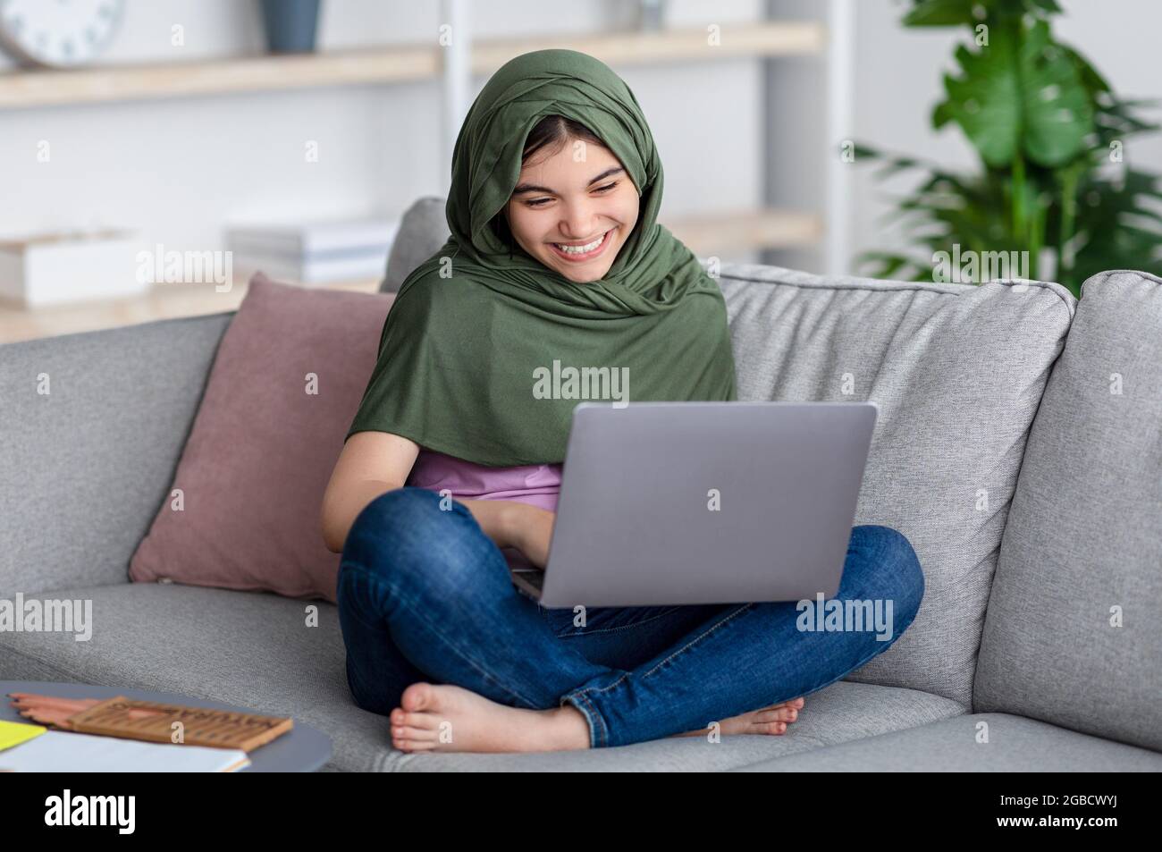 Concetto di apprendimento remoto. Felice ragazza teen musulmana in hijab studiare online, seduto sul divano con il computer portatile a casa Foto Stock
