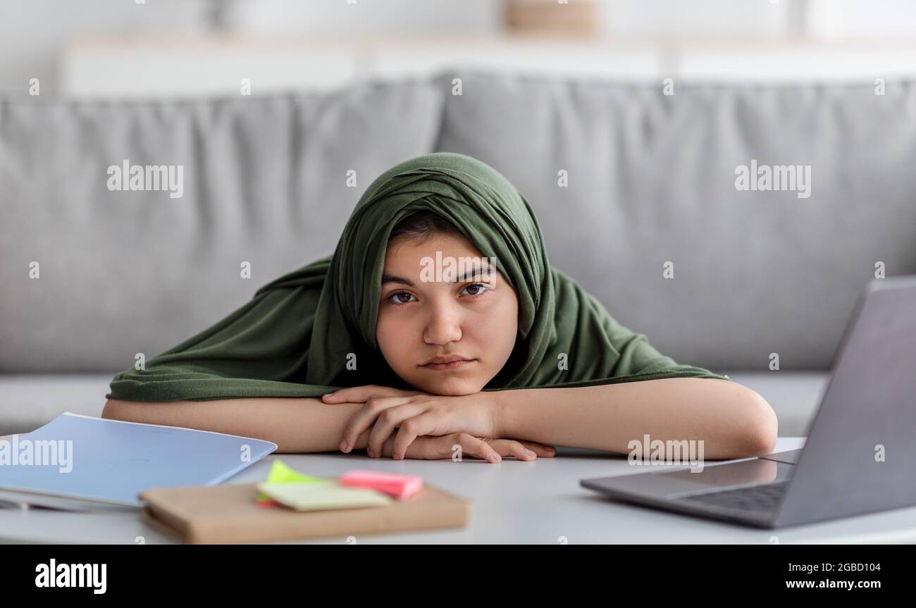 Ragazza teen musulmana in hijab sdraiata sul tavolo con il laptop e materiali di studio, avendo una lezione remota opaca, l'apprendimento online Foto Stock