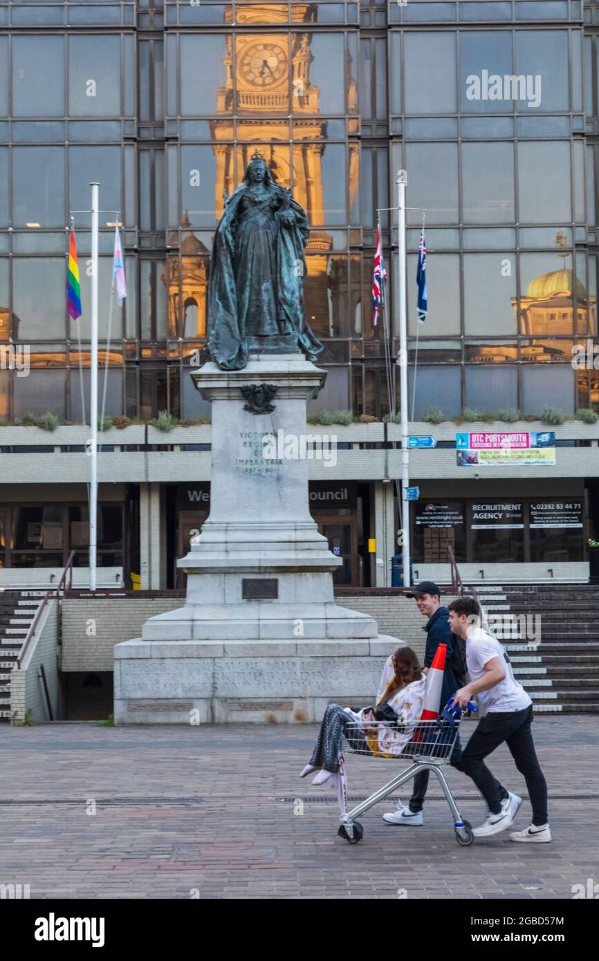 Inghilterra, Hampshire, Portsmouth, Guildhall Square, studenti universitari che spingono il supermercato Shopping Trolly di fronte alla statua della Regina Victoria Foto Stock