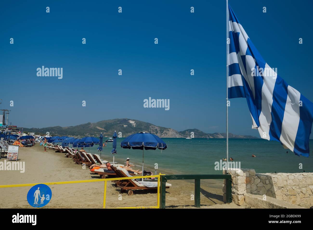 Zante, Grecia terreno mediterraneo organizzato spiaggia con bandiera greca e bagnanti. Giorno soleggiato vista del lungomare di Laganas con mare calmo e lettini con ombrelloni sotto il cielo blu. Foto Stock