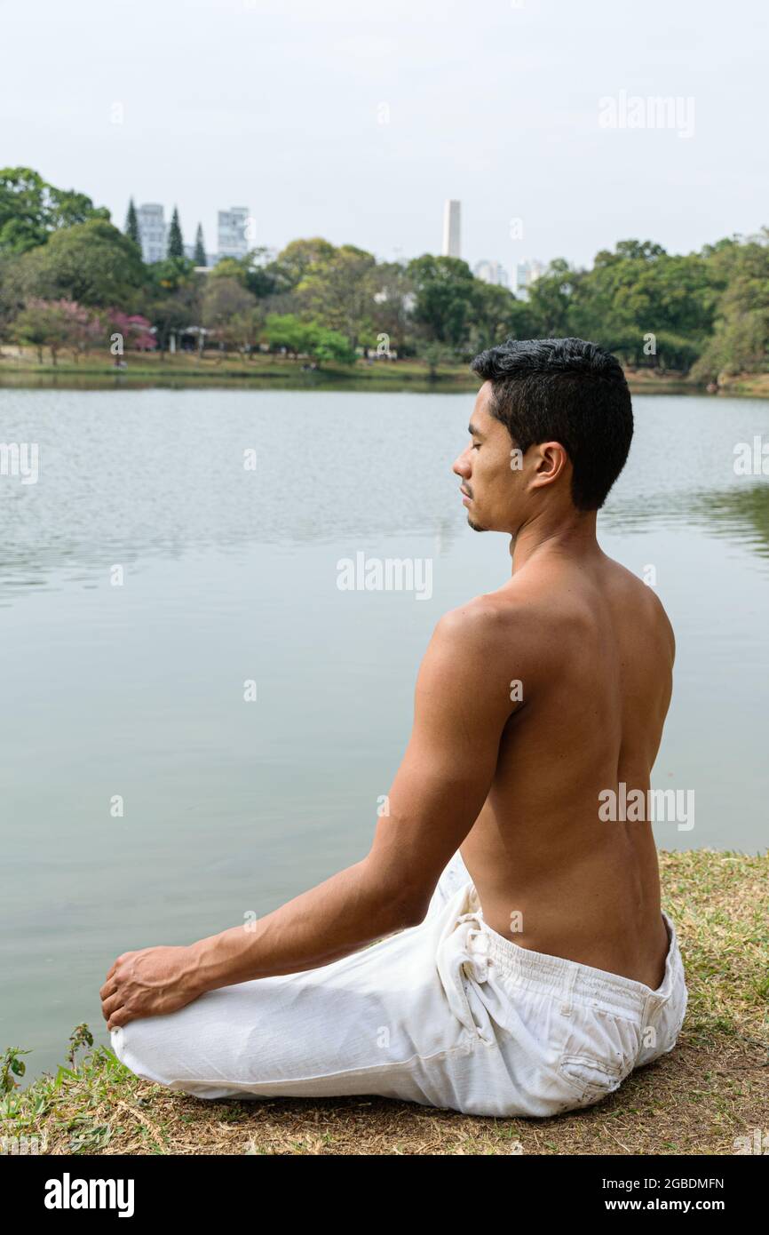 Giovane brasiliano seduto guardando il lago in posizione di meditazione (foto verticale). Foto Stock