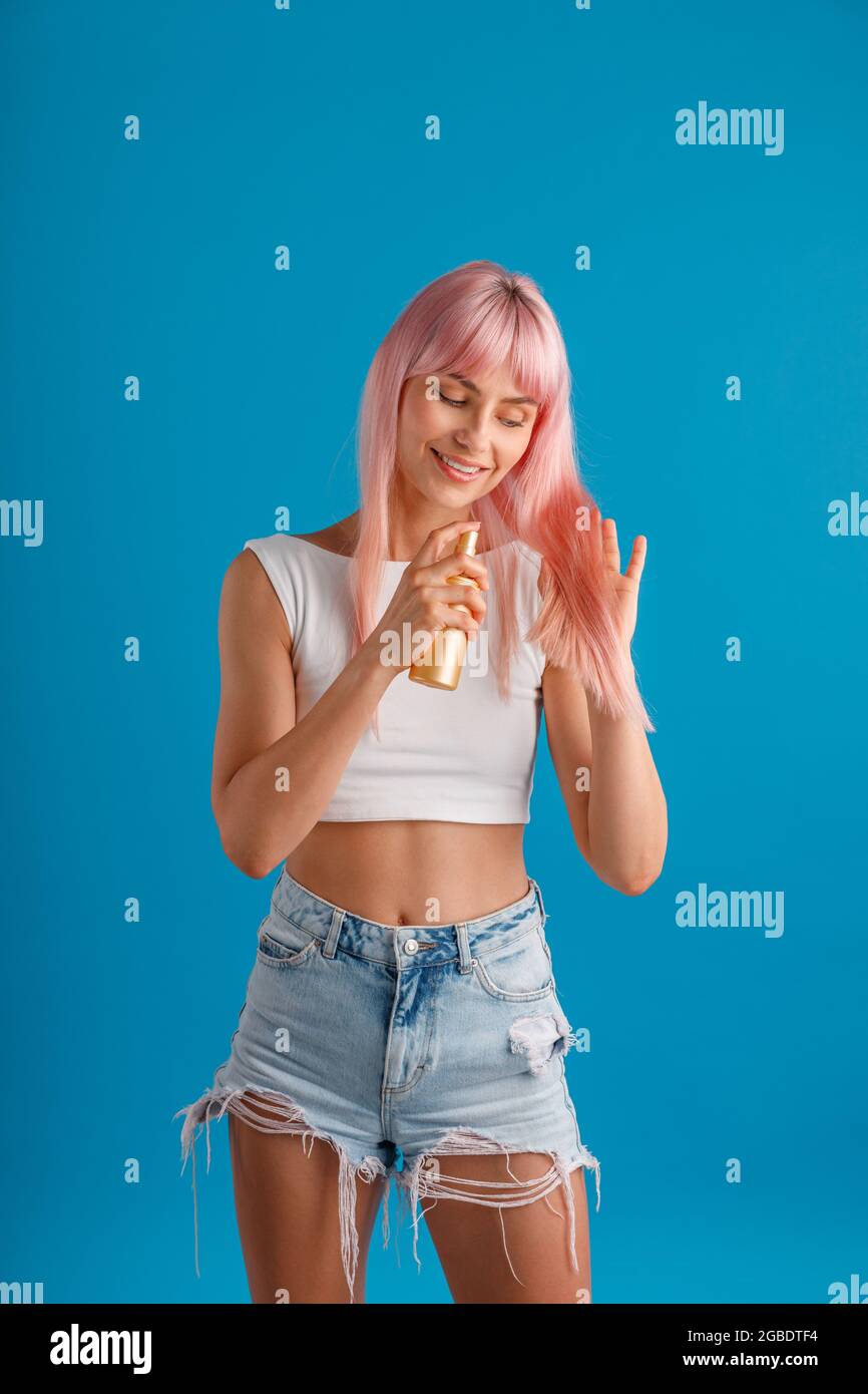 Allegra giovane donna sorridente e applicando spray idratante ai suoi bei capelli lisci rosa tinti mentre si trova isolata sopra studio blu Foto Stock