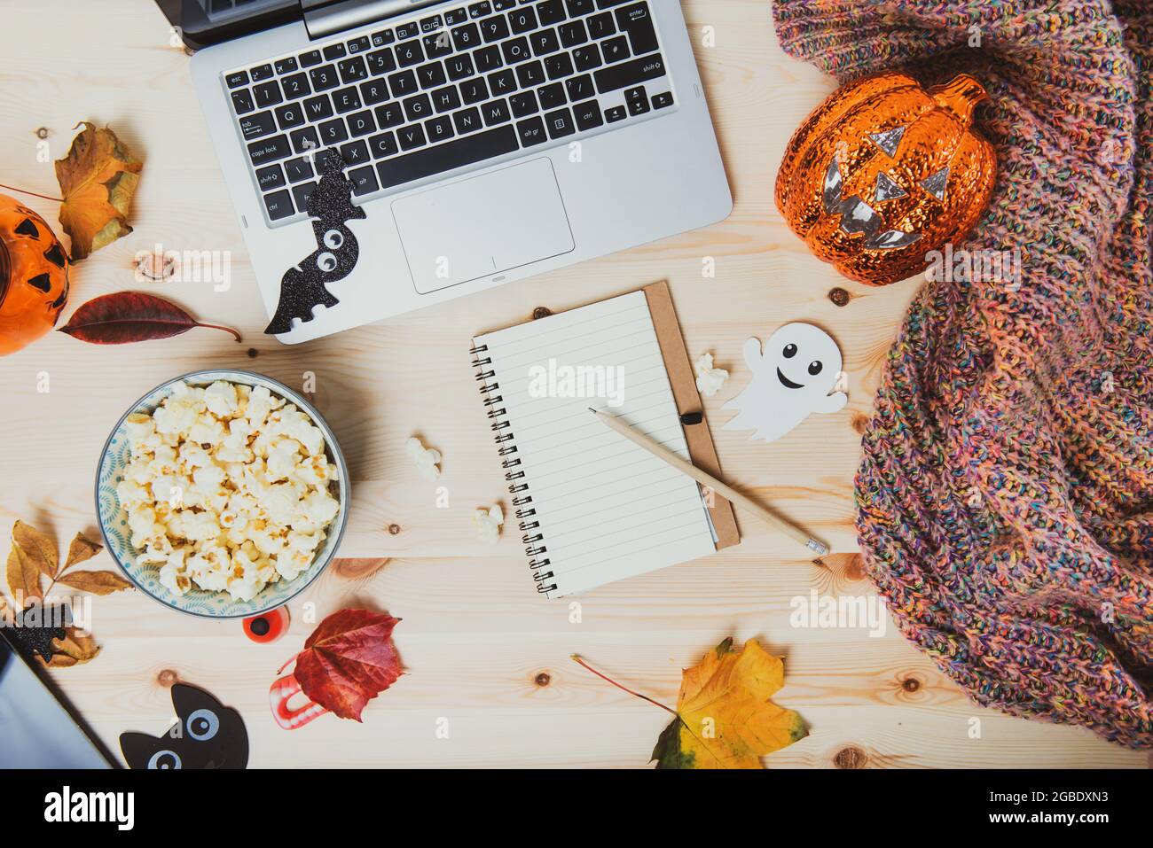 Notebook vuoto con vista dall'alto su un tavolo di legno con computer portatile, popcorn, decorazioni di Halloween, zucca con illuminazione, foglie d'autunno e plaid caldo. Elenco di pianificazione dei film Foto Stock
