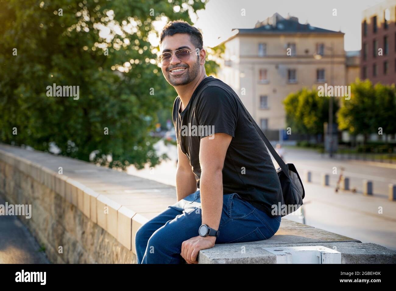 Giovane arabo indiano o ispanico in camicia nera con una borsa a tracolla e occhiali da sole seduti su una parete sorridente mentre si guarda la fotocamera contro bl Foto Stock