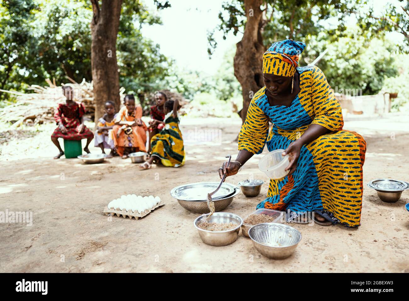 In questa immagine, una madre africana nera vestito di colore sta preparando un pasto per la sua famiglia, con le sue figlie in attesa sullo sfondo Foto Stock