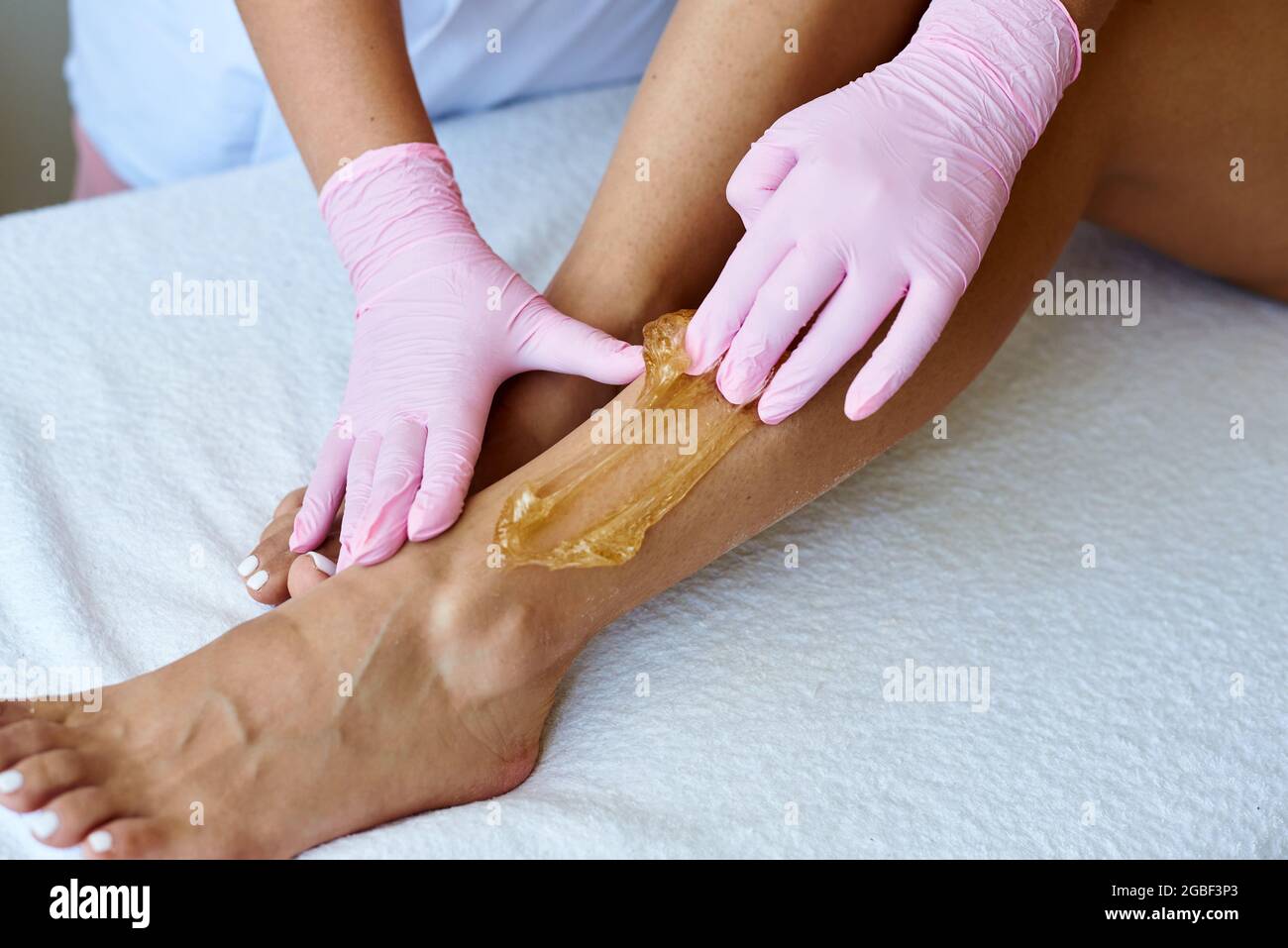 Il giovane e grazioso estetista applica la pasta di zucchero alla gamba del cliente. Epilazione con shugaring. Estetista depilante giovane donna. Foto Stock