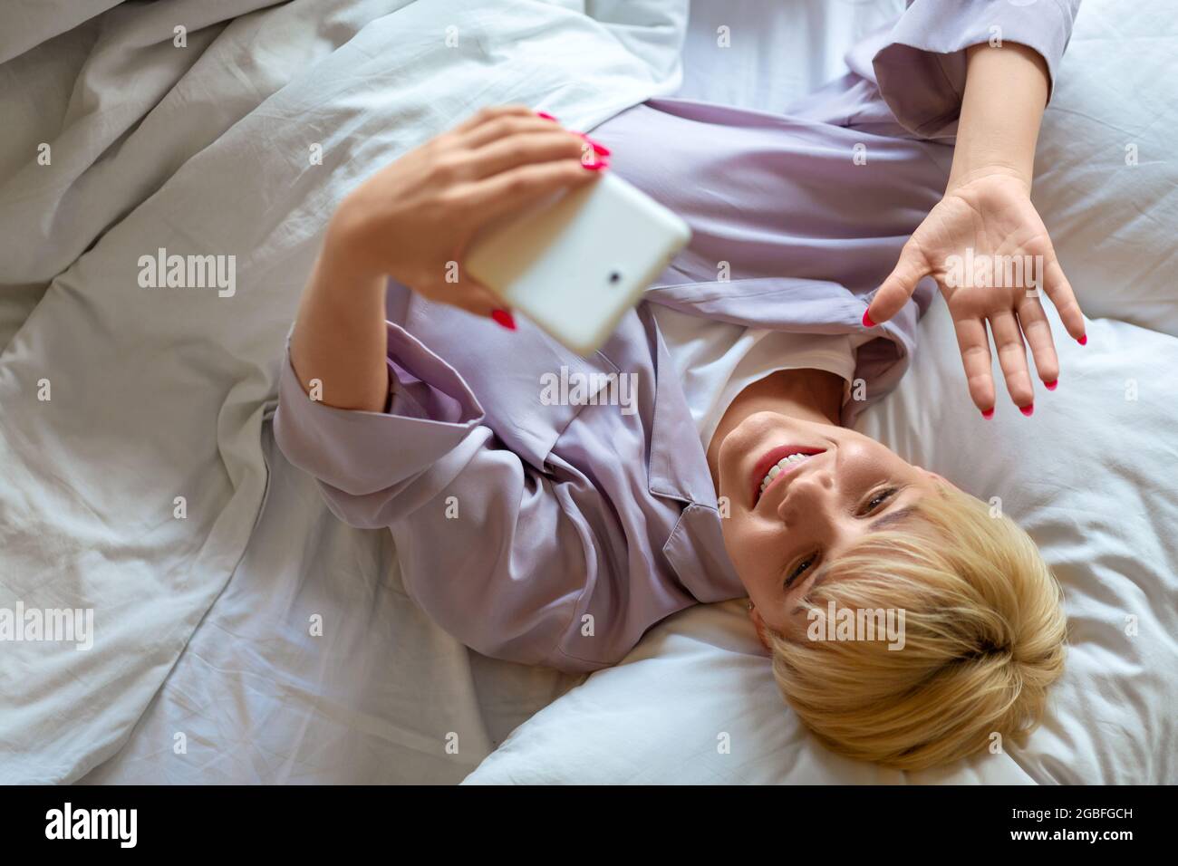 Giovane donna a letto durante la videochiamata online. Incontro online con gli amici. Lei agita la mano e dice ciao dopo il risveglio. Foto Stock