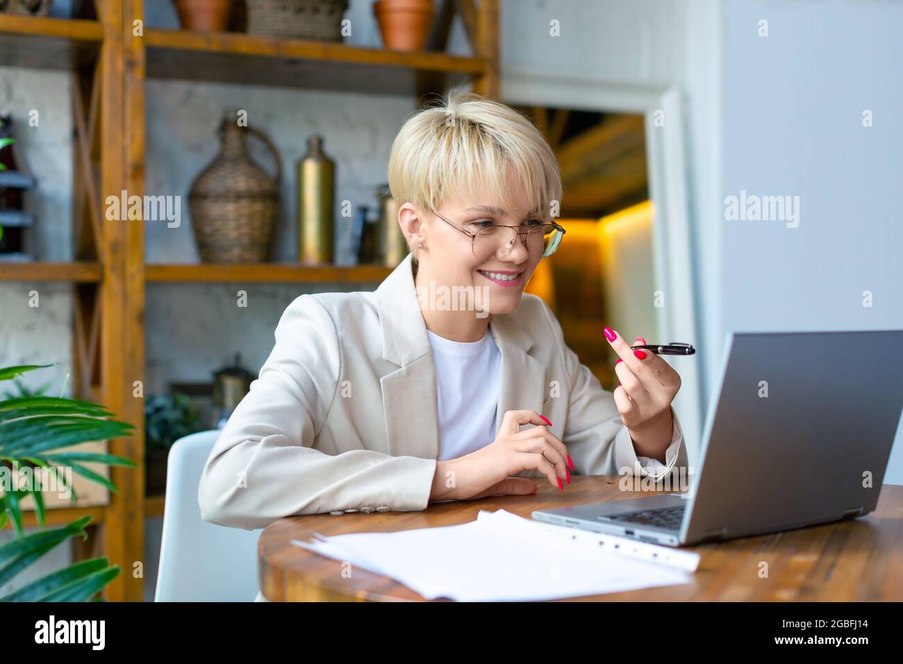 Foto verticale di una giovane bionda con occhiali davanti a un laptop. Sorride durante una videochiamata online con i colleghi e punta con una penna Foto Stock