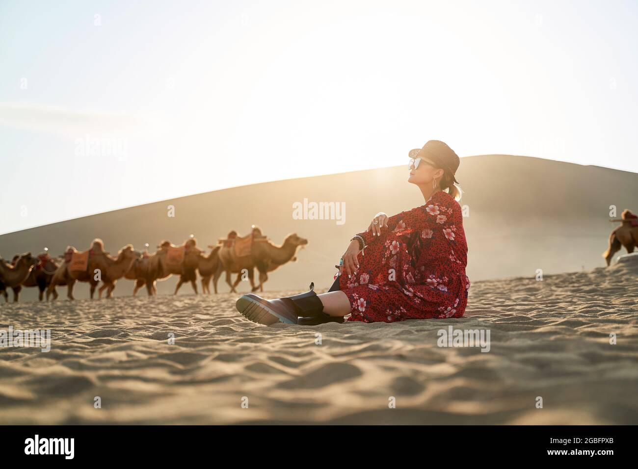 donna asiatica in abito rosso seduta nel deserto guardando la vista con caravan di cammelli e enorme duna di sabbia sullo sfondo Foto Stock