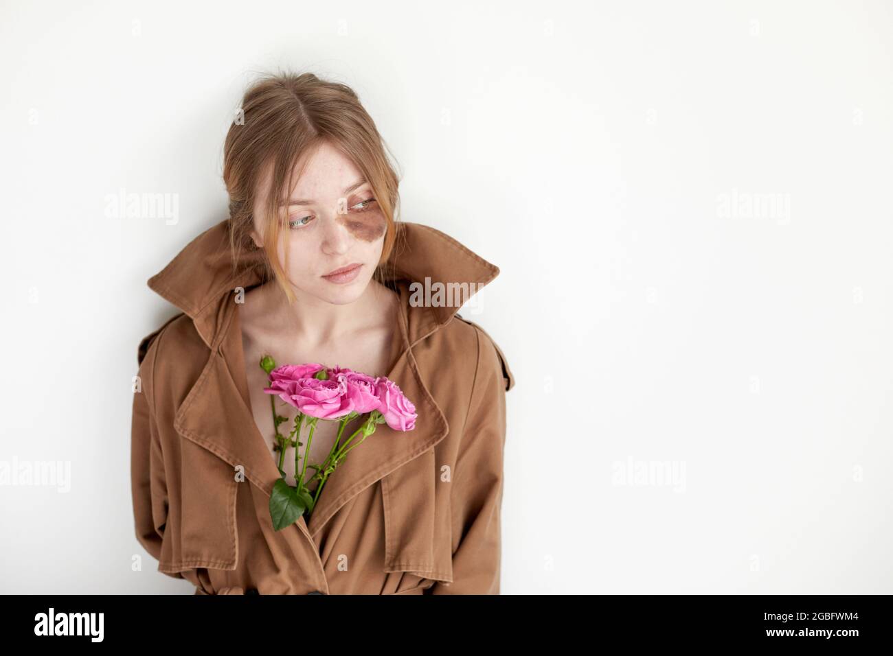 Giovane modello femminile con punto scuro sotto l'occhio vestito con cappotto marrone con fiori in fiore brillante in piedi contro parete bianca Foto Stock