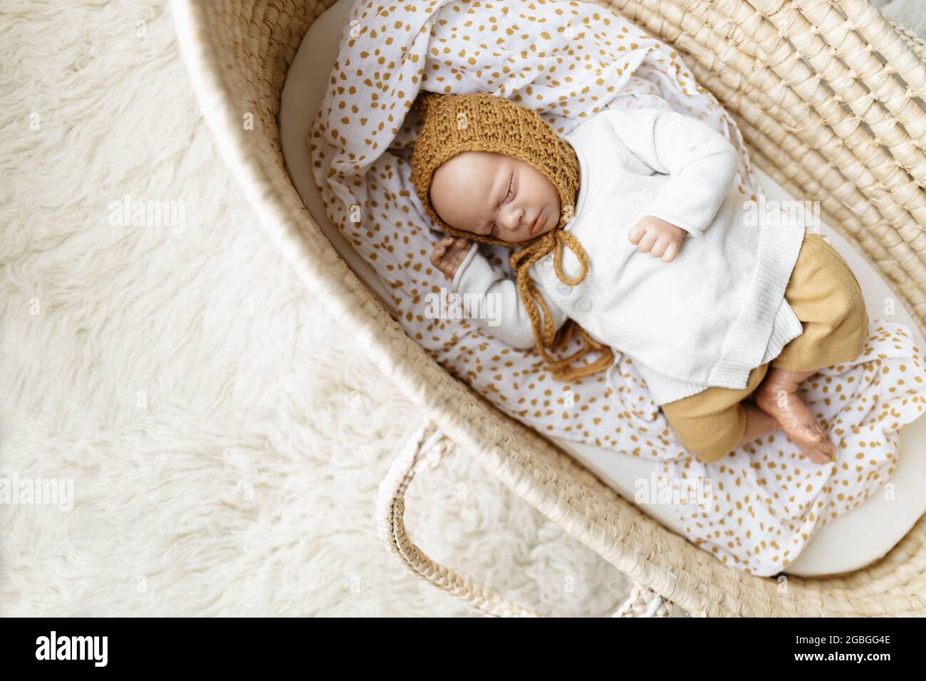 Un bambino neonato neonato giocattolo vestito con un cappello sul cofano color senape e un cardigan bianco sdraiato in una crema di fagotto moses basket Foto Stock