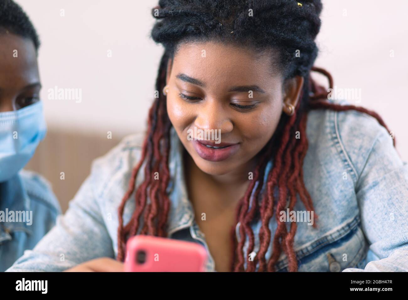 Una donna afroamericana si siede con un telefono rosa nelle mani. Un'altra donna coetanei sopra la sua spalla. Foto Stock