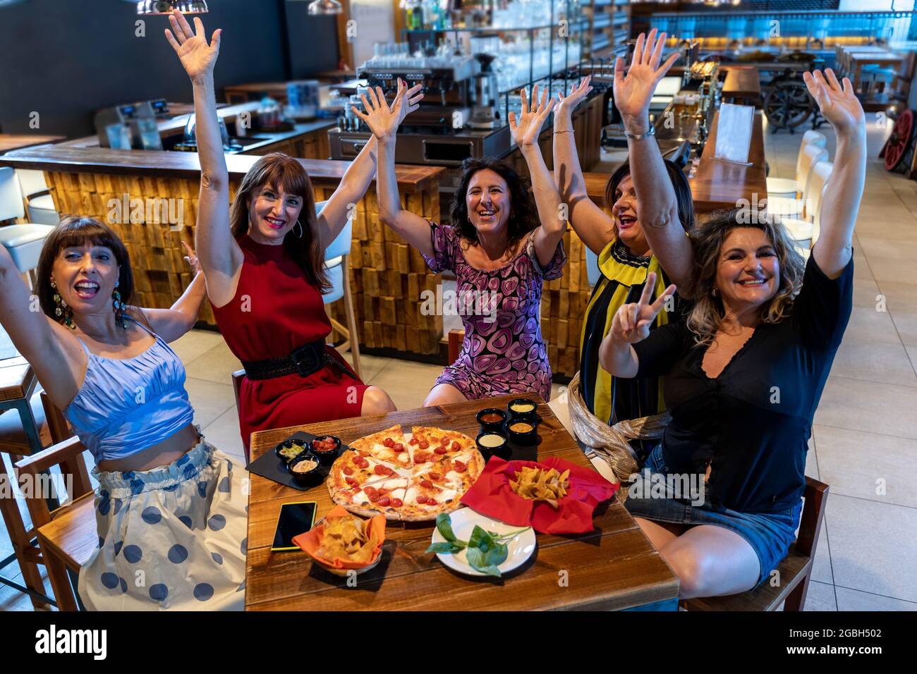 gruppo di donne mature sedute in un pub alzano le mani mentre mangiano la pizza e bevono la birra Foto Stock