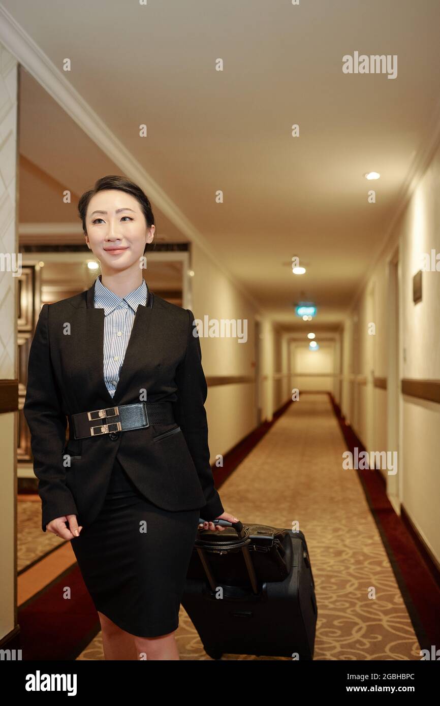 Contetn attraente giovane donna asiatica in formalwear essendo in viaggio d'affari a piedi con i bagagli lungo il corridoio dell'hotel Foto Stock