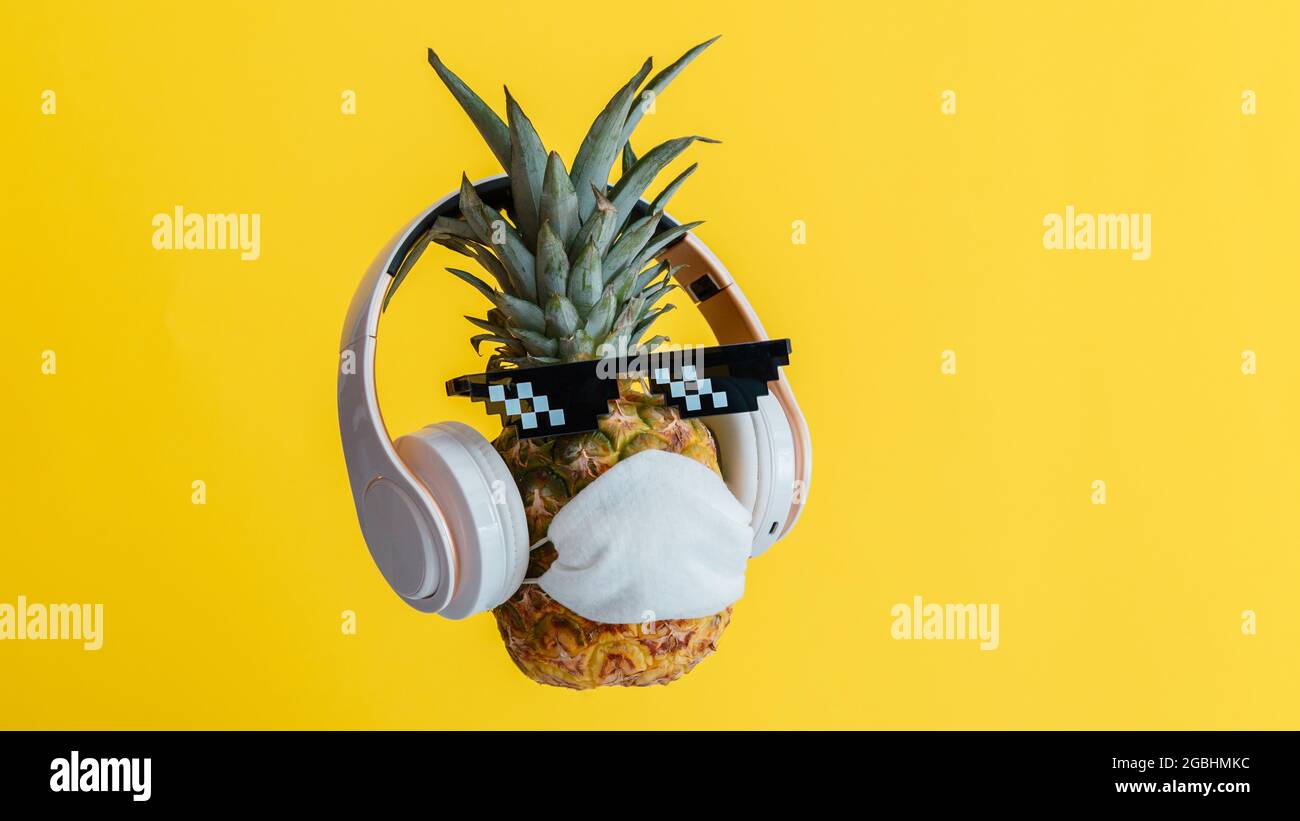 Creativo divertente ananas viso indossare occhiali cuffie e maschera medica di protezione. Riposante faccia di ananas levitating sul colore giallo estate Foto Stock