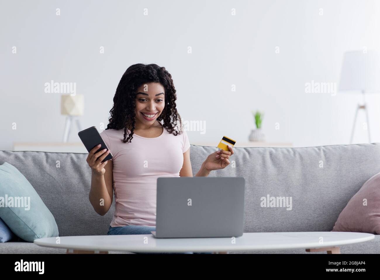 Giovane donna sorridente che usa lo smartphone che acquista online con carta di credito sul divano quando si rilassa in soggiorno a casa Foto Stock