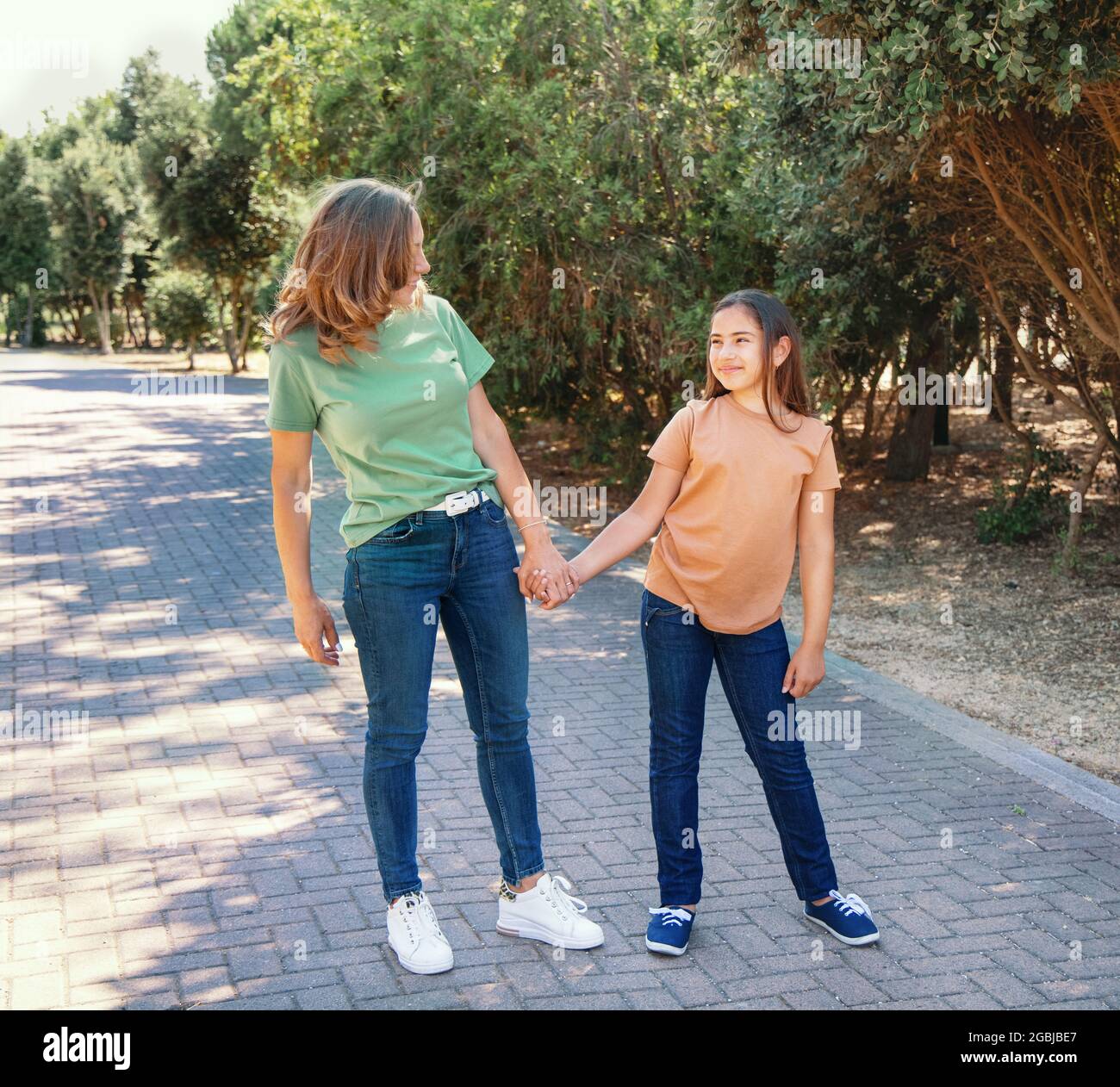 Madre e figlia che camminano nel parco. Una donna di mezza età e una ragazza di capretto che si tengono le mani l'un l'altro, facendo il contatto dell'occhio e indossando t- verde e arancione Foto Stock