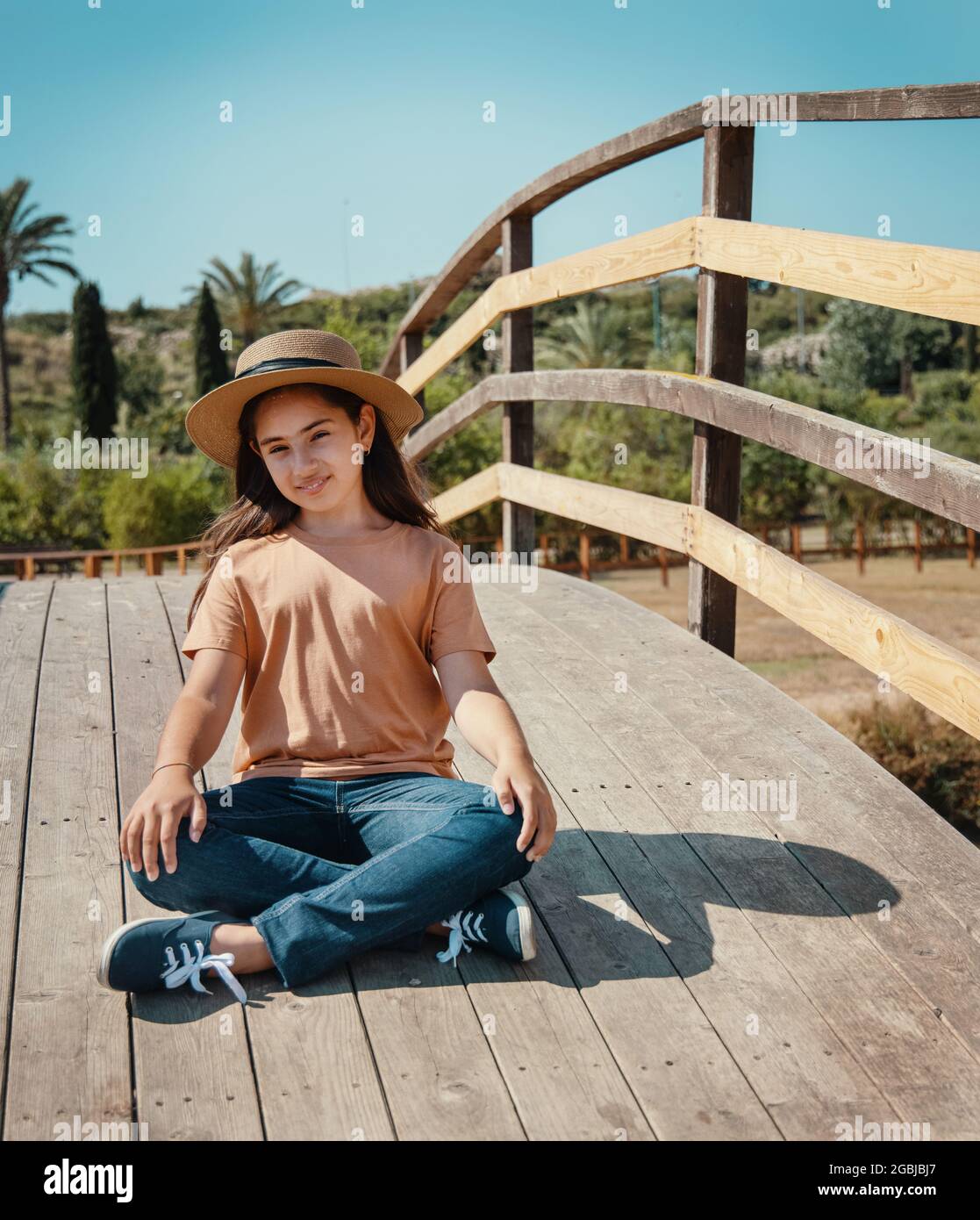 Una bambina sorridente seduto croce zampe sul ponte di legno nel parco in giornata di sole indossando cappello di paglia, t-shirt arancione e jeans e guardando dritto Foto Stock
