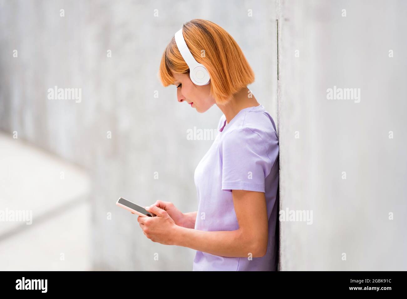 Attraente donna rossa che ascolta la musica sulle cuffie stereo che naviga sul suo telefono cellulare per le colonne sonore mentre si appoggia contro una wa bianca Foto Stock