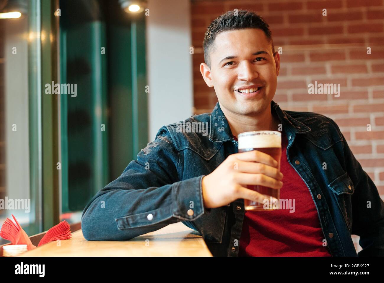 Felice attraente uomo ispanico gustando una birra fredda in un pub o bar alzando il suo bicchiere e dando alla macchina fotografica un caldo sorriso amichevole Foto Stock
