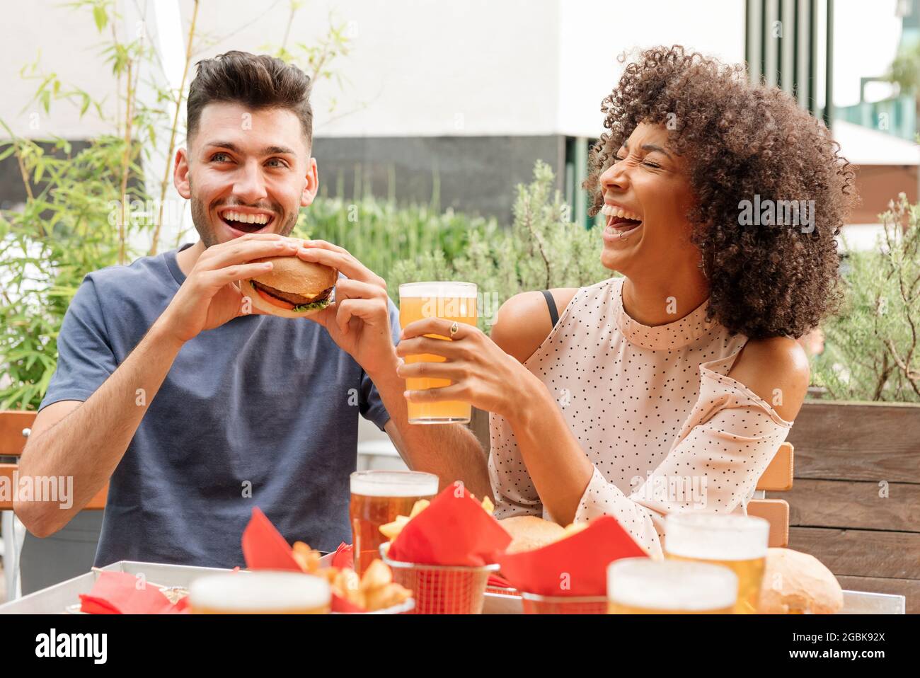 Felice affamata coppia multiculturale gustare hamburger e birre fredde in un ristorante all'aperto o un pub ridendo felice a una buona scherzata Foto Stock