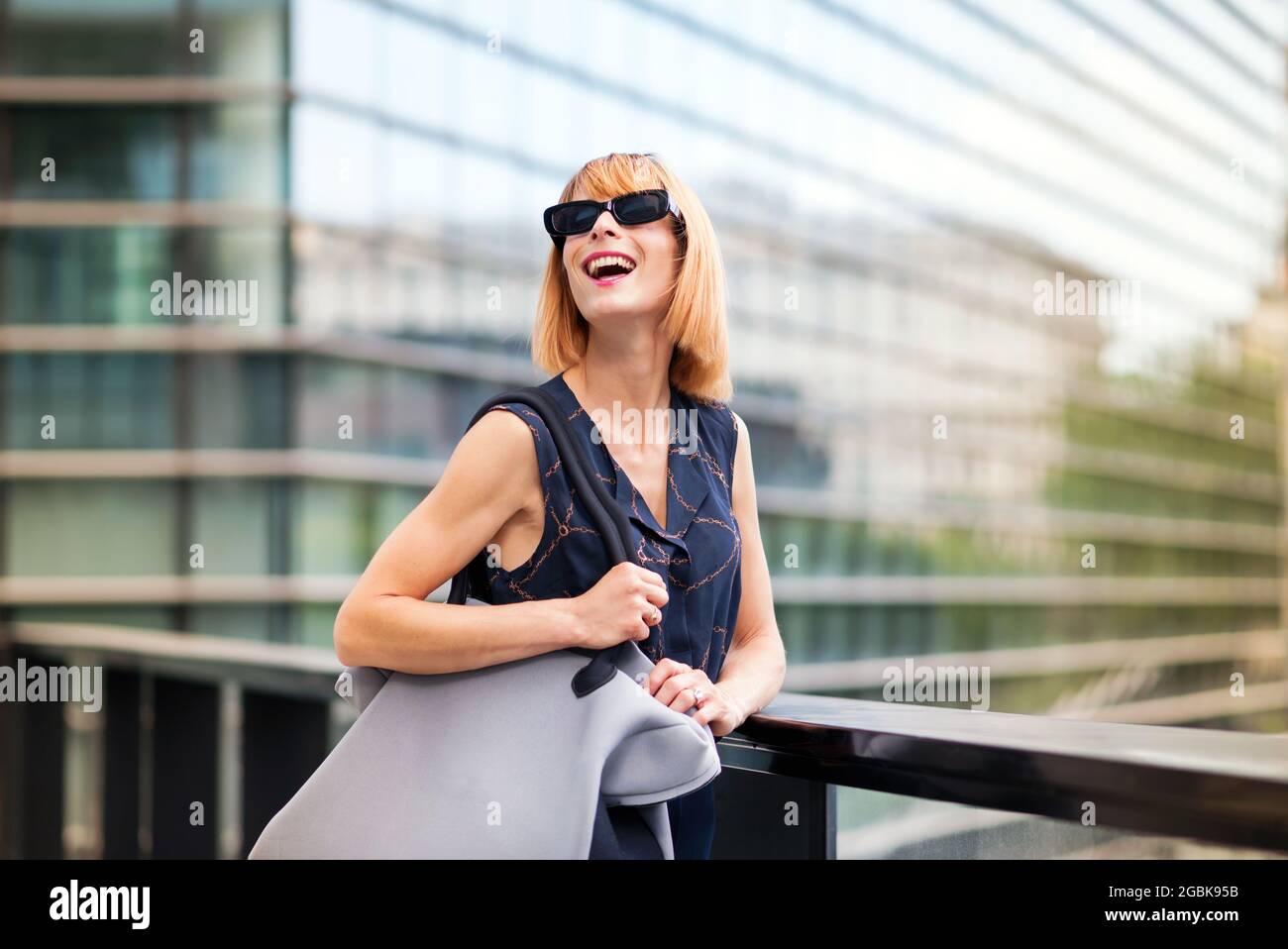 Vivace donna trendy in moderni occhiali da sole in piedi contro la ringhiera di un passaggio in una città ridendo felice mentre guarda verso il cielo con vetro Foto Stock