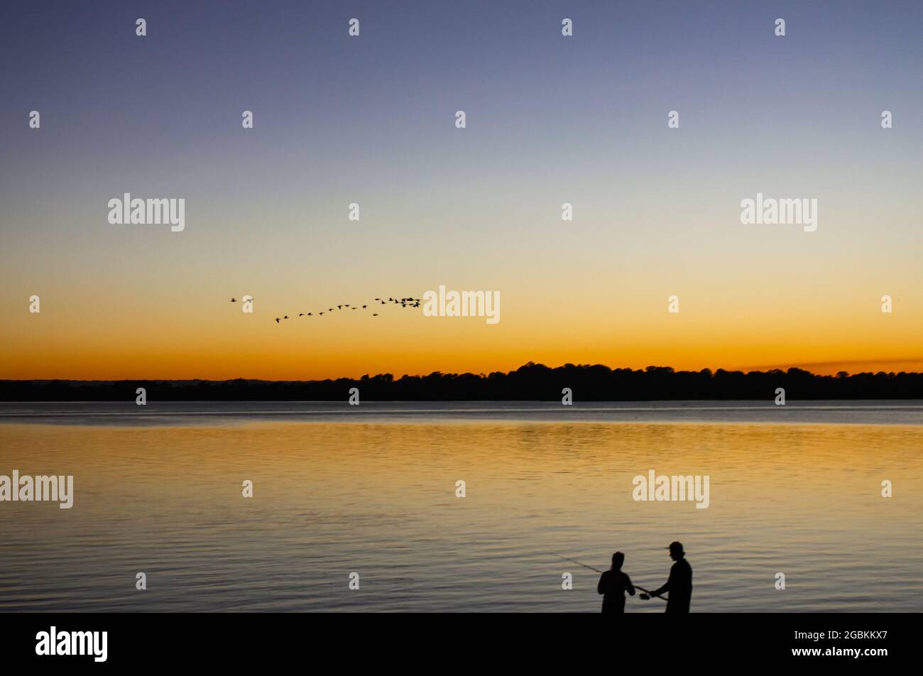 Sagome di pesca uomo e donna con tramonto che si riflette in acqua e gregge di uccelli che volano in formazione in alto Foto Stock